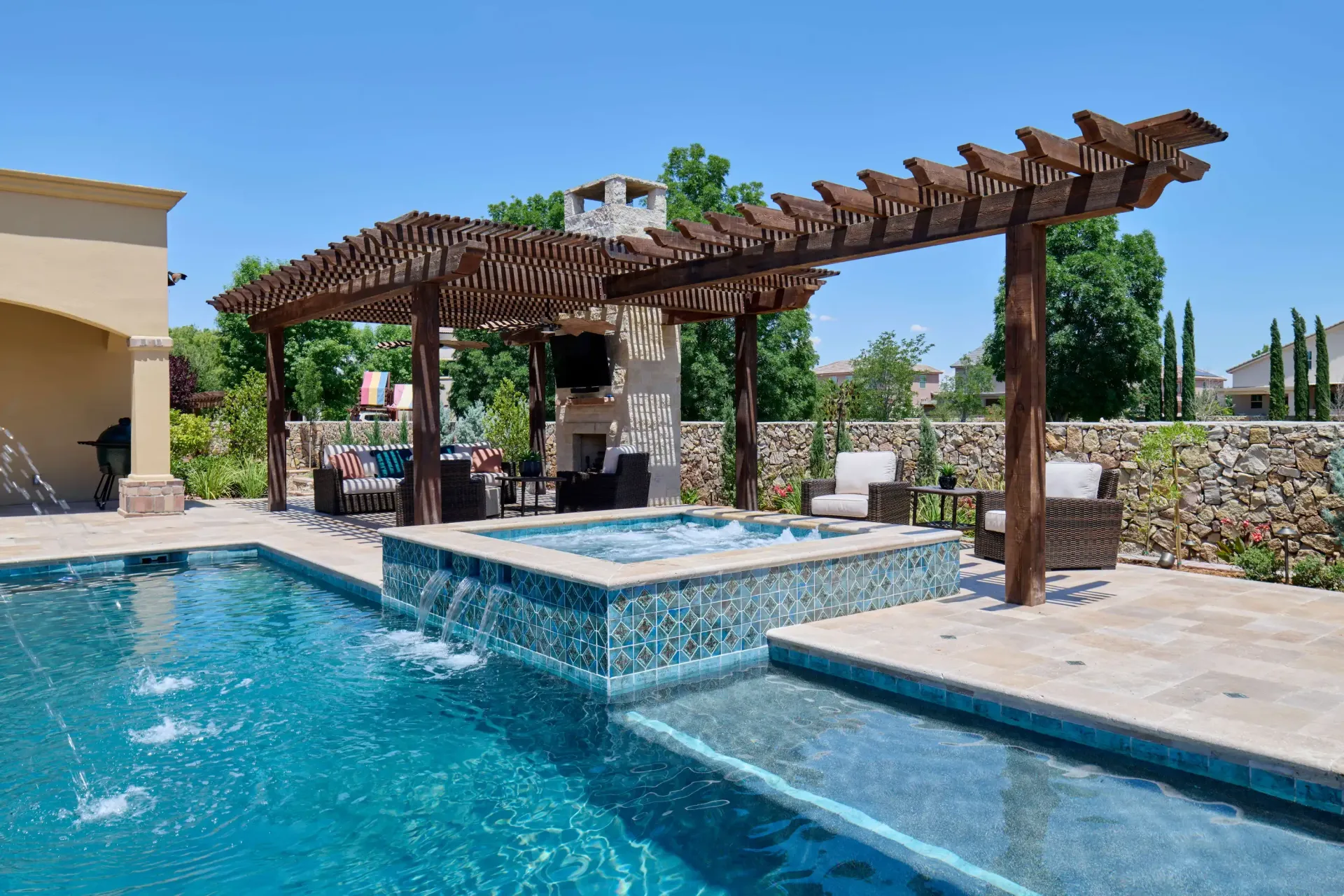 Pool area with pergola, hot tub, and fireplace under a clear, blue sky.
