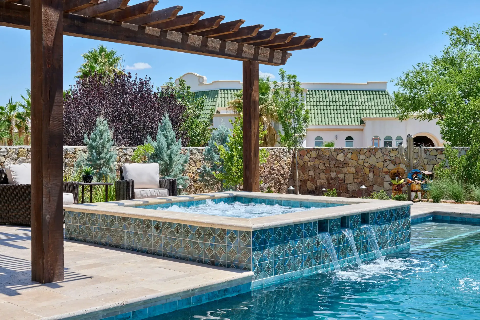A backyard with a pool, spa, and pergola. The spa has a waterfall, blue water, and is surrounded by stone.