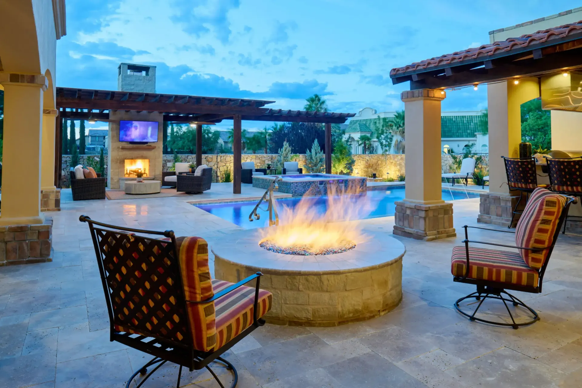 Outdoor patio with fire pit, pool, seating, fireplace, and television. Evening scene with blue sky.
