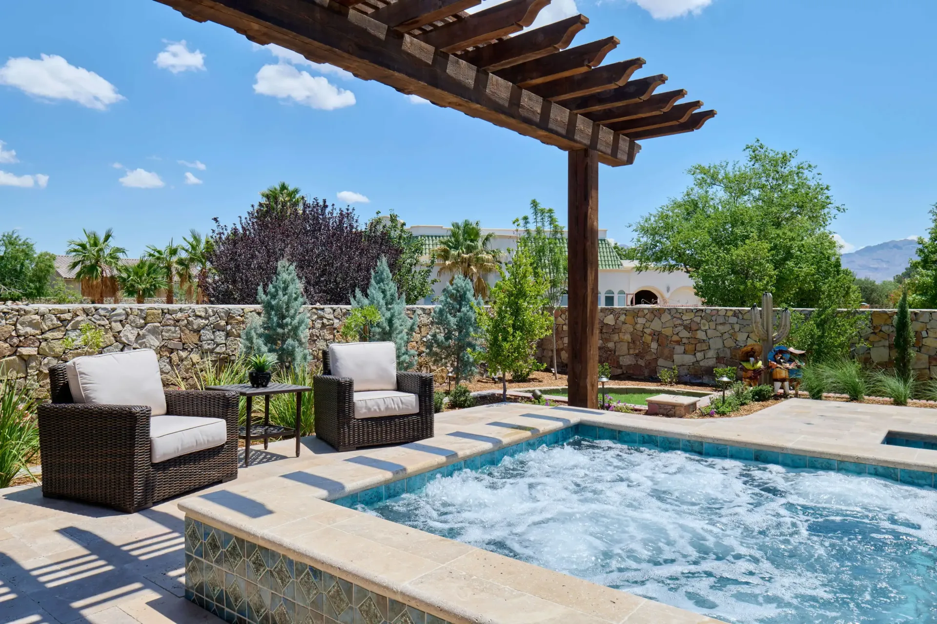 Two wicker chairs under a pergola beside a bubbling spa, in a sunny outdoor setting.