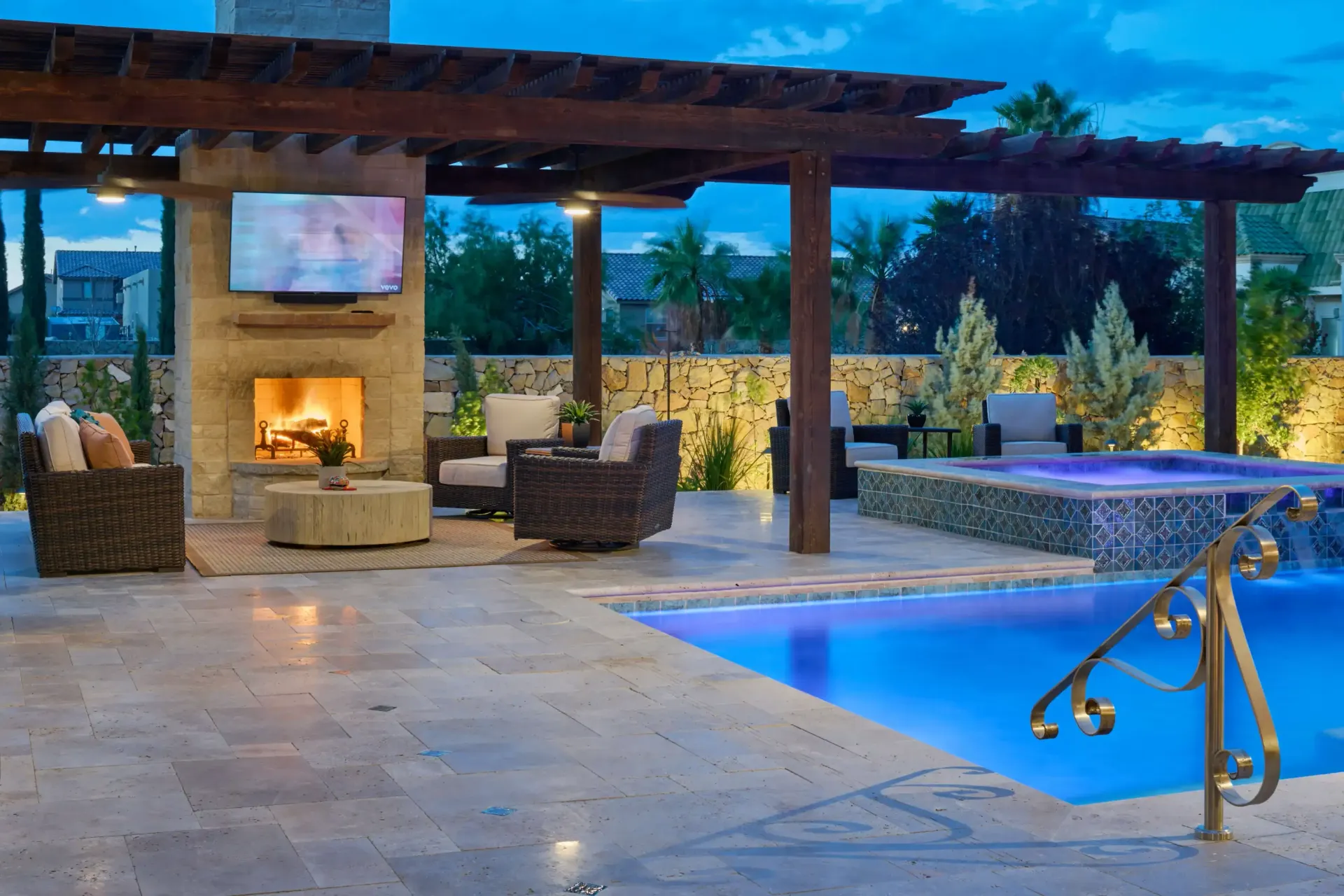 Outdoor patio with fireplace, TV, seating area, pool, and pergola, illuminated at dusk.