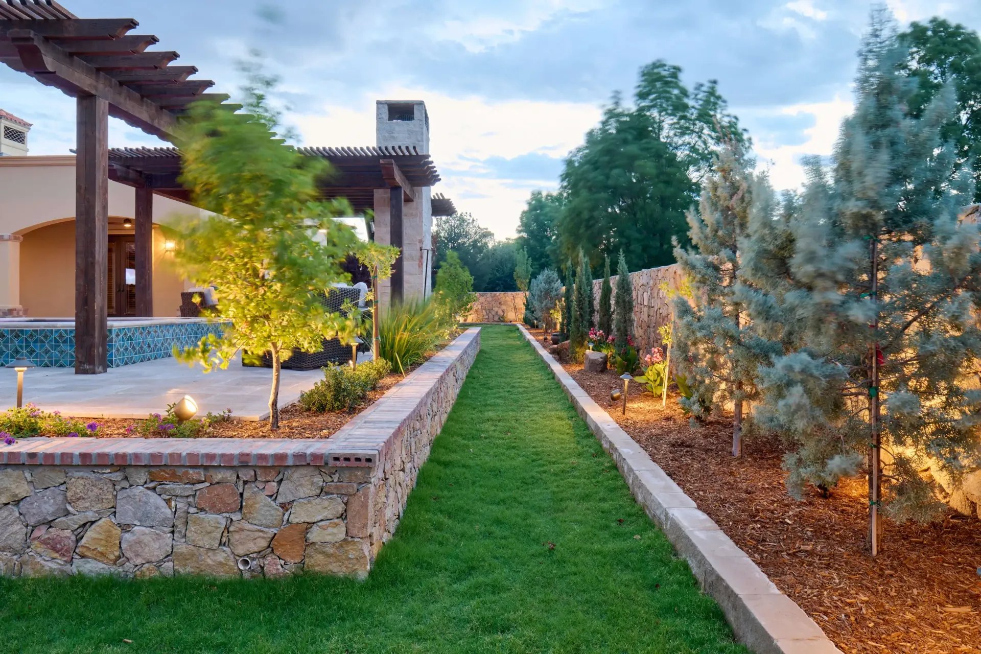 Pathway lined with grass, trees, and stone walls, leading to a home with a wooden pergola.