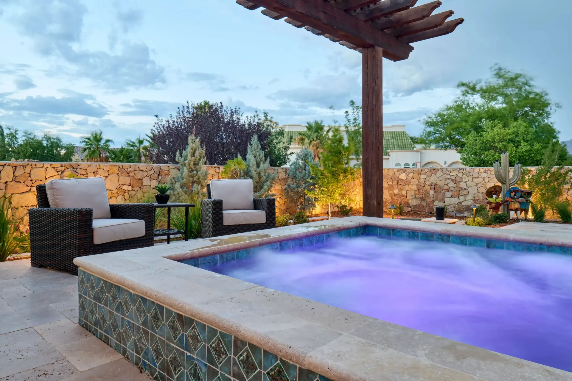 Outdoor spa with lounge chairs under a pergola, set against a stone wall with plants and a cloudy sky.