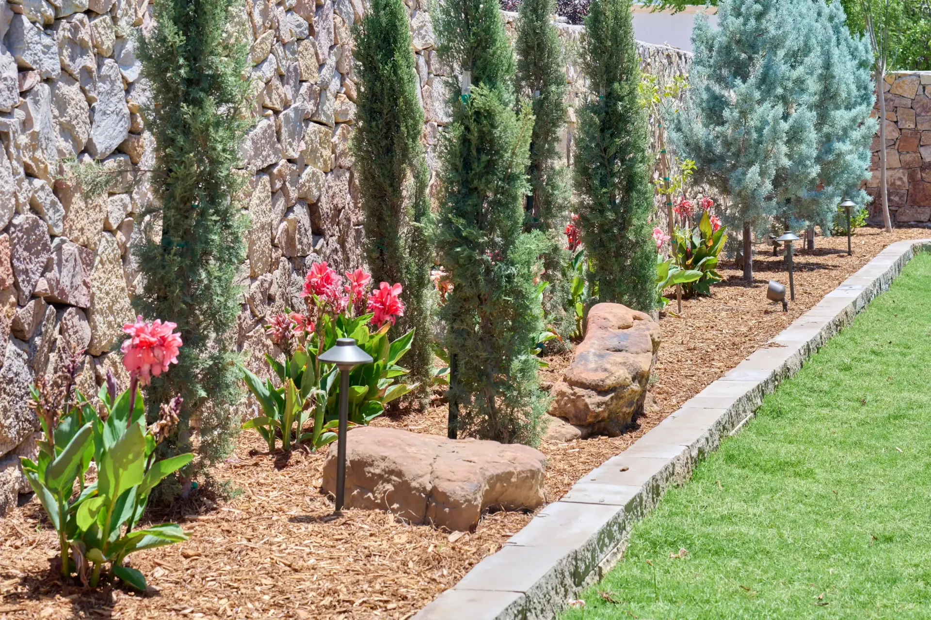 Landscaped garden with tall evergreen trees, pink flowers, and a stone wall.