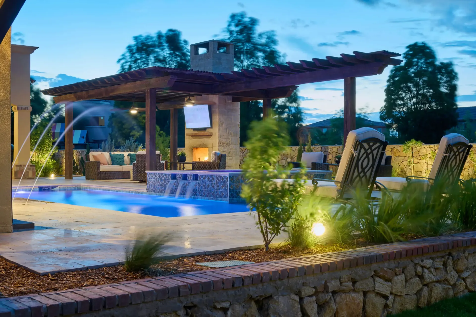 Poolside patio with pool, pergola, fireplace, and water fountain, at dusk.