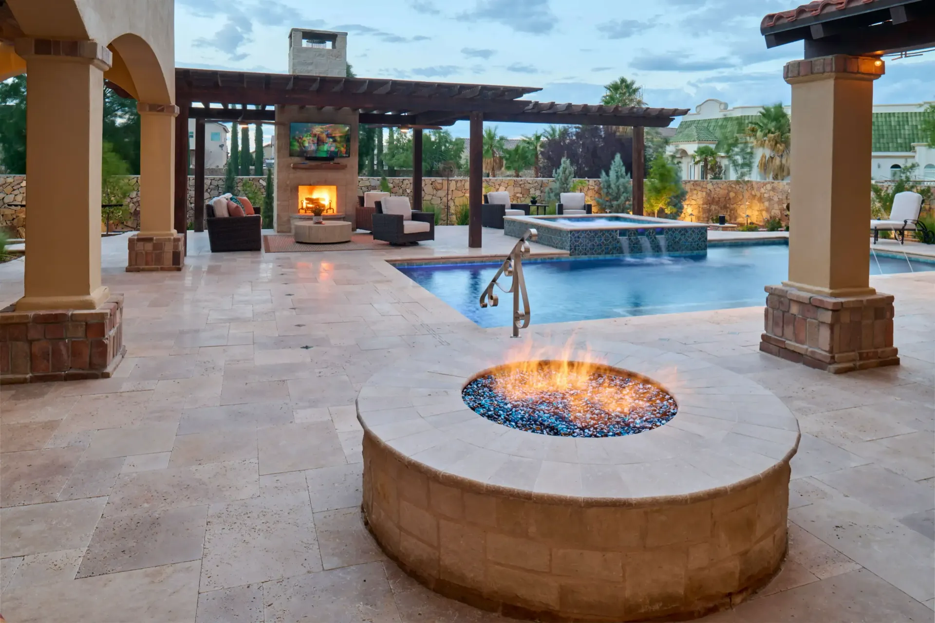 Outdoor patio with pool, fireplace, and fire pit. Blue flames in fire pit, pool water reflects sky, pergola overhead.