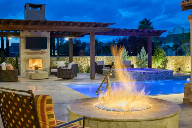 Outdoor patio with fire pit, pool, hot tub, fireplace, and seating under a pergola at dusk.