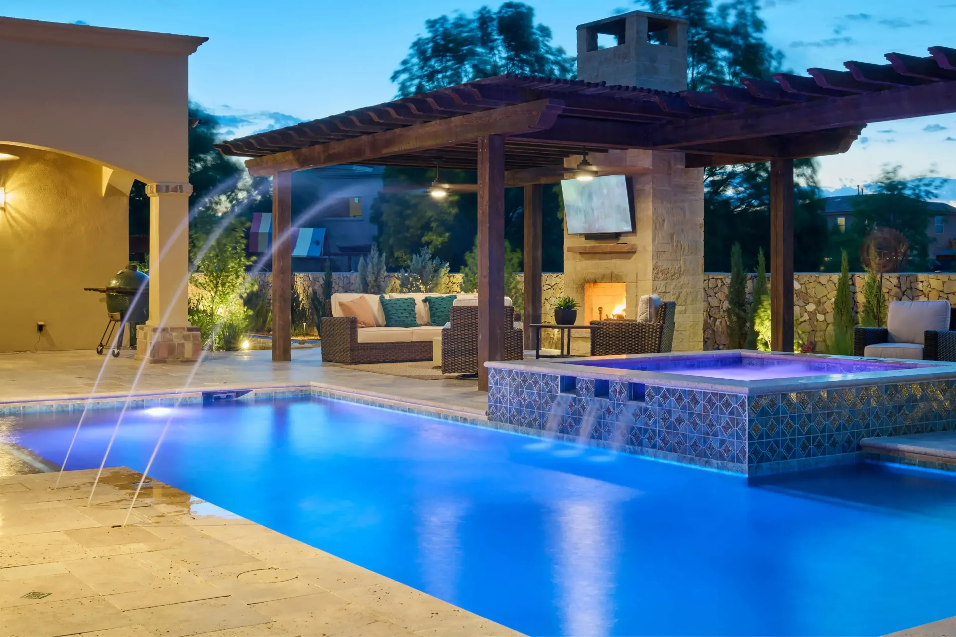 Outdoor pool area with blue-lit water, jacuzzi, fireplace, seating under a pergola. Evening light.