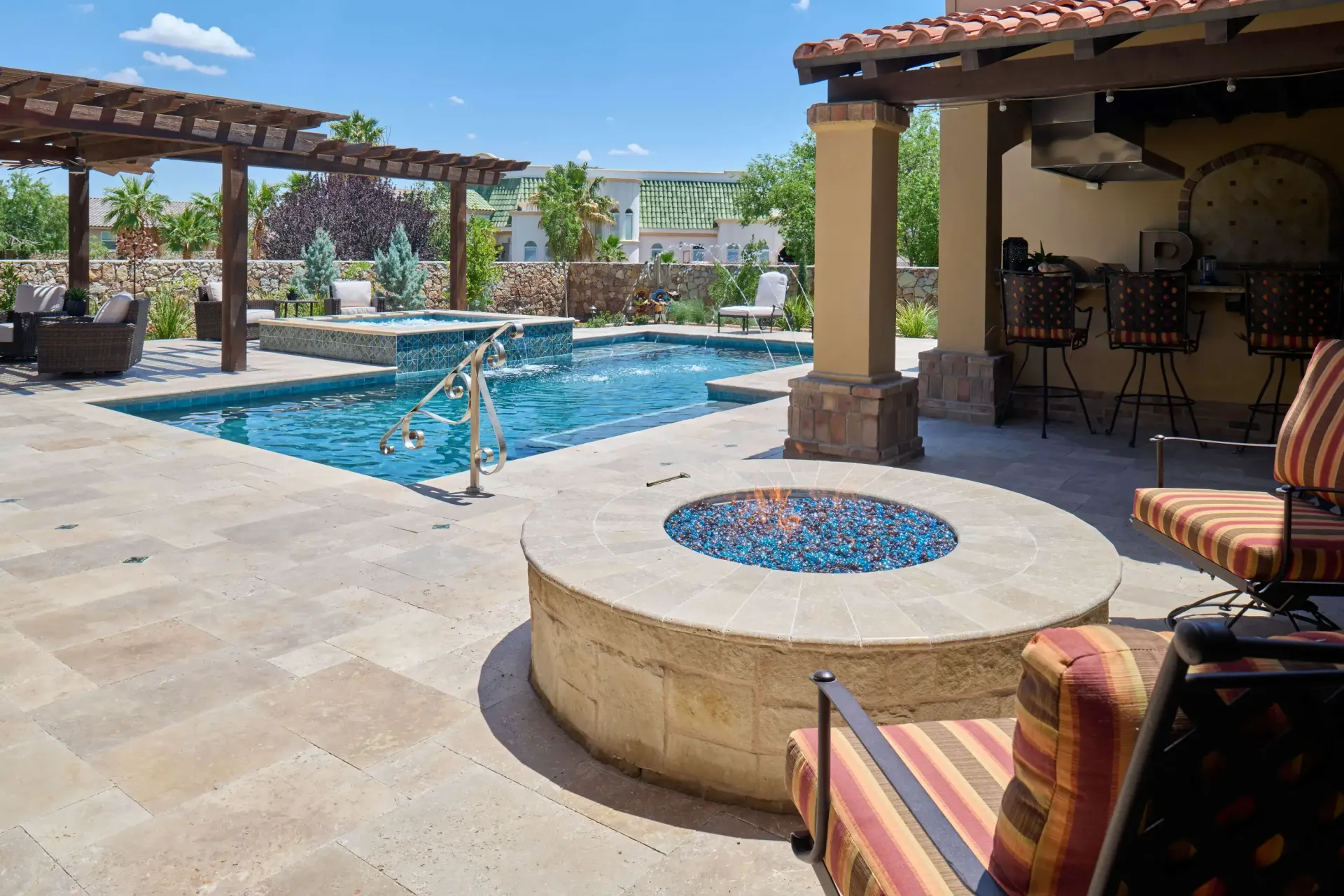 Patio with pool, fire pit, and bar seating. Blue pool water, stone pavers, and striped chairs.