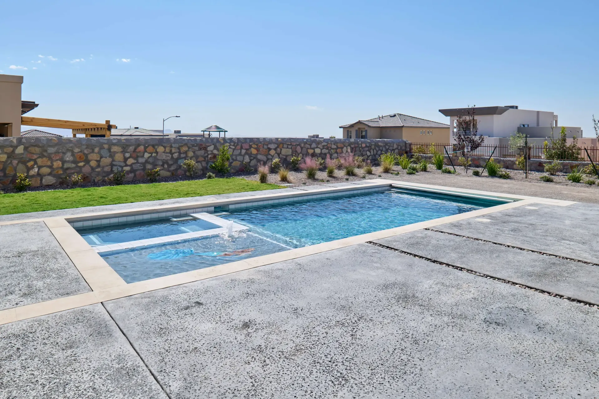 Rectangular pool with attached jacuzzi. Concrete patio, stone wall, and houses in the background under blue sky.