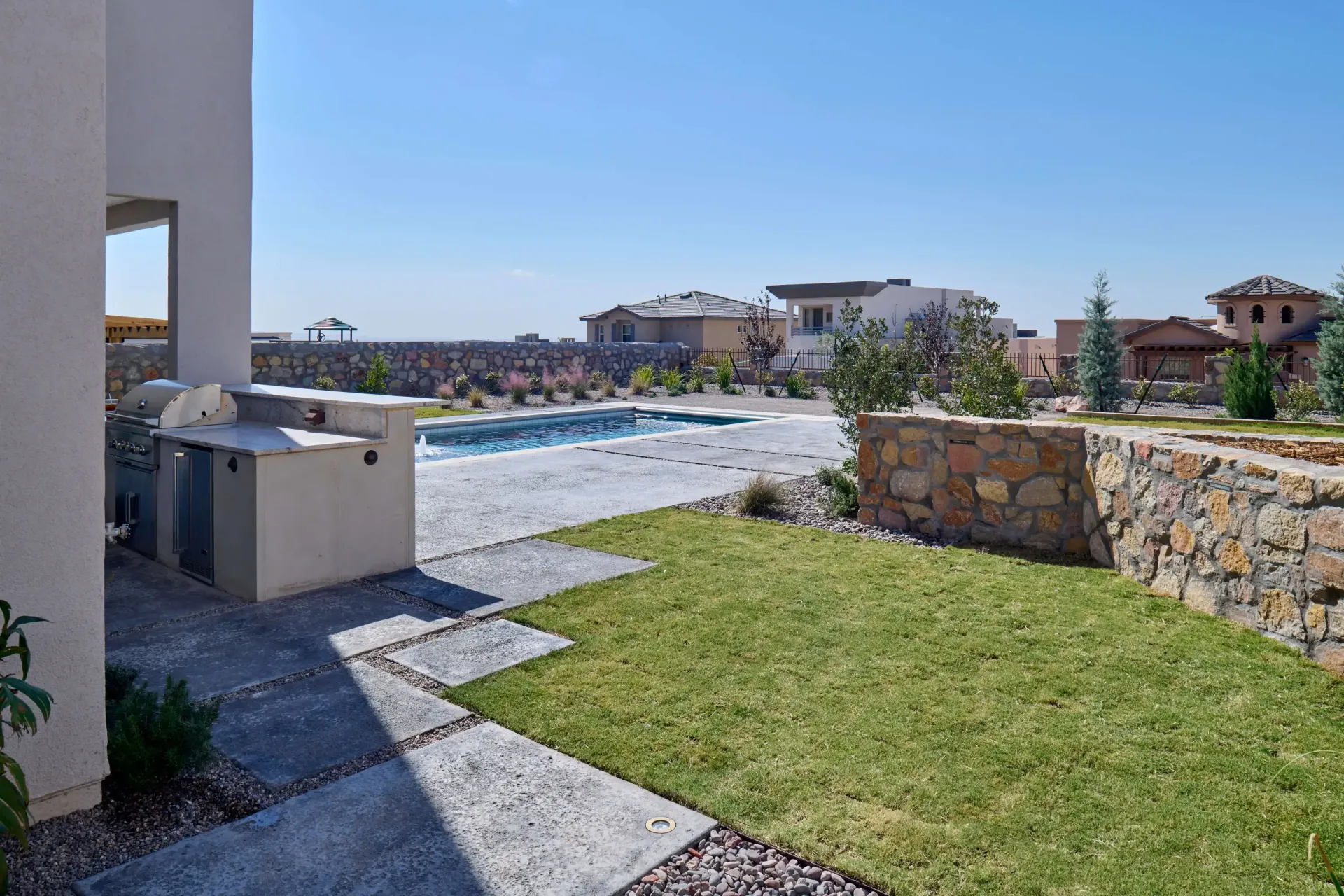 Backyard with built-in grill, pool, stone walls, and green lawn under a bright blue sky.