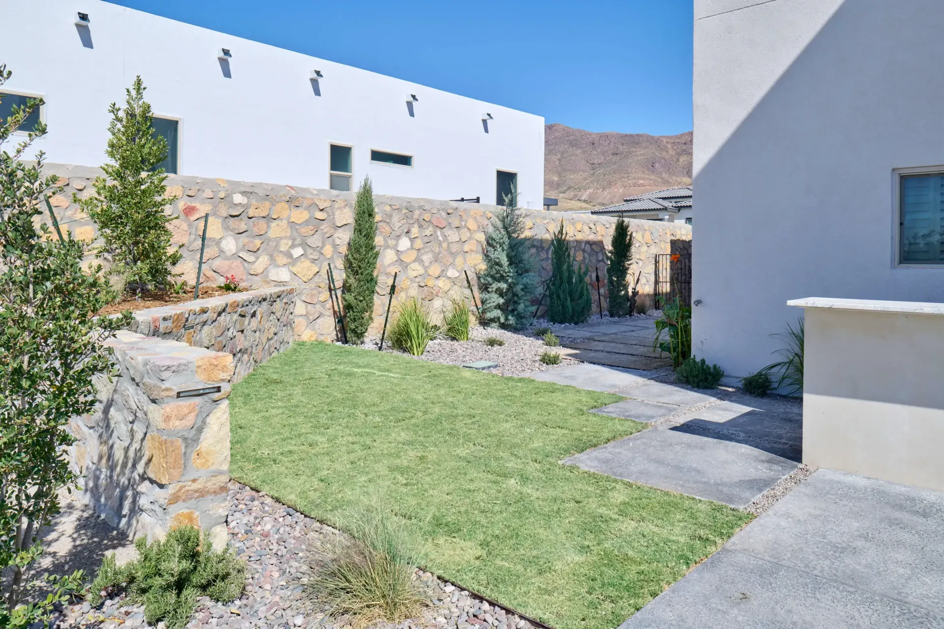 A small backyard with a patch of green grass, stone walls, and white buildings in the background.