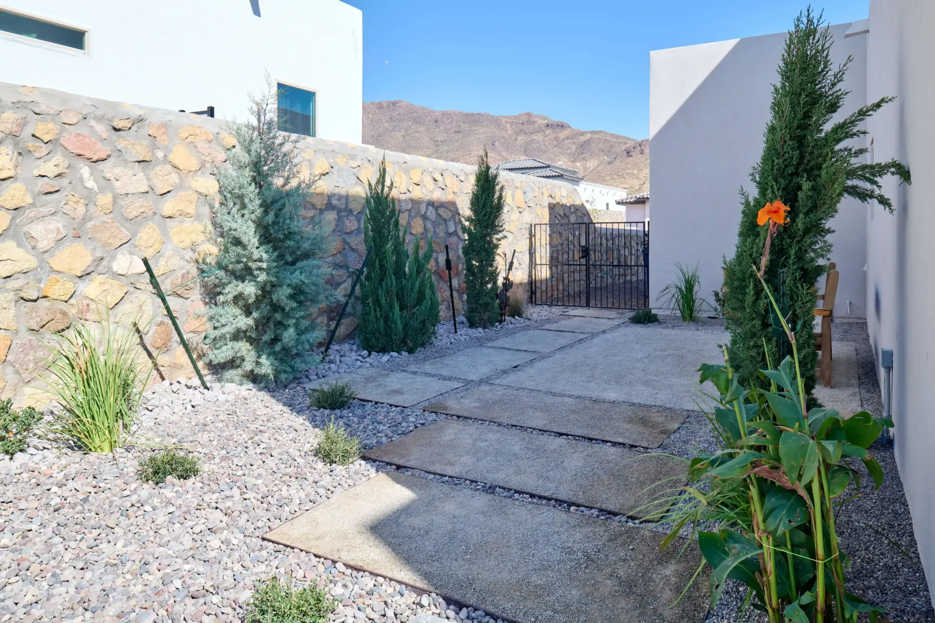 Pathway through a gravel yard with stone walls and evergreen trees against a mountain backdrop.