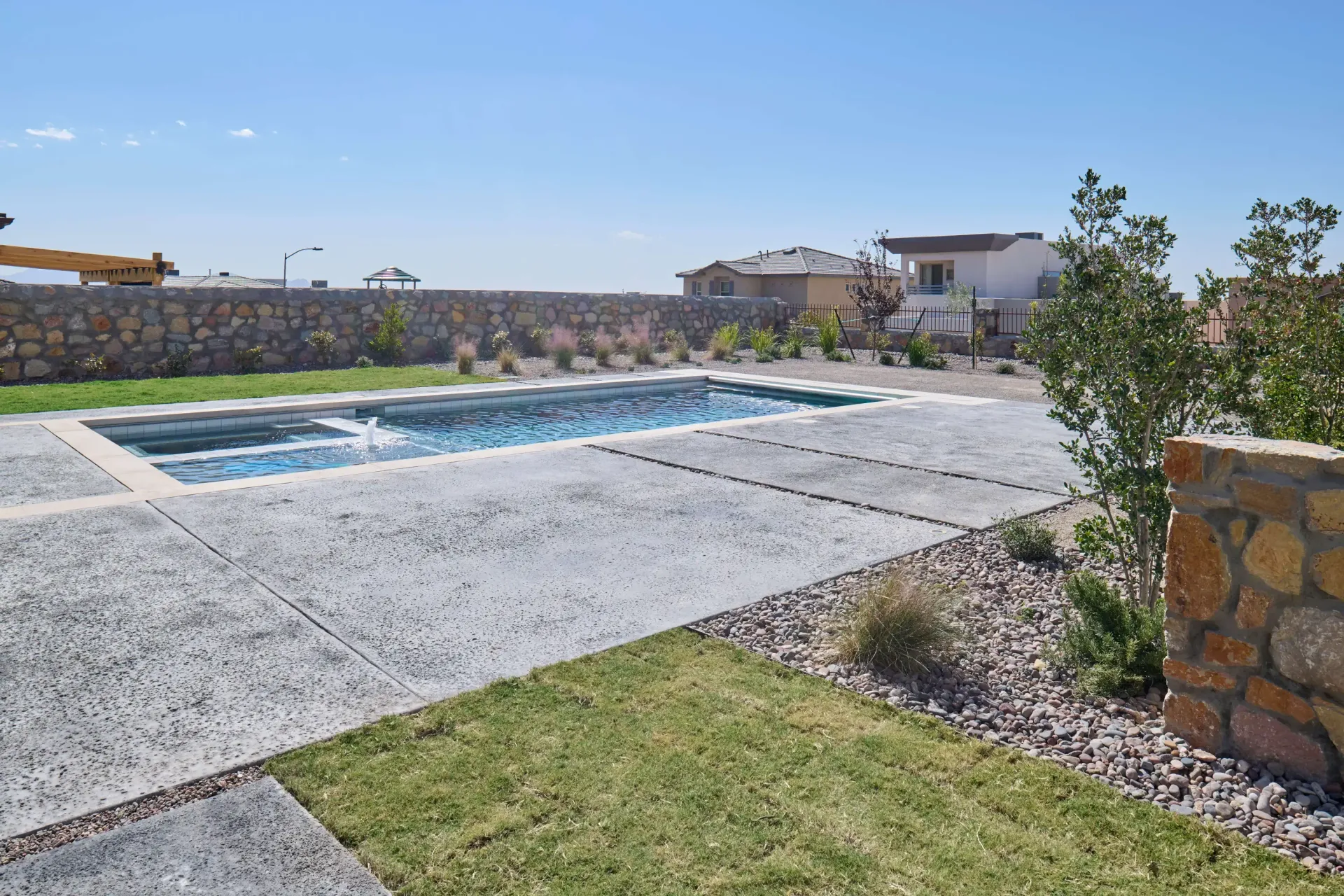 Pool surrounded by concrete and grass, set against a stone wall and buildings under a blue sky.
