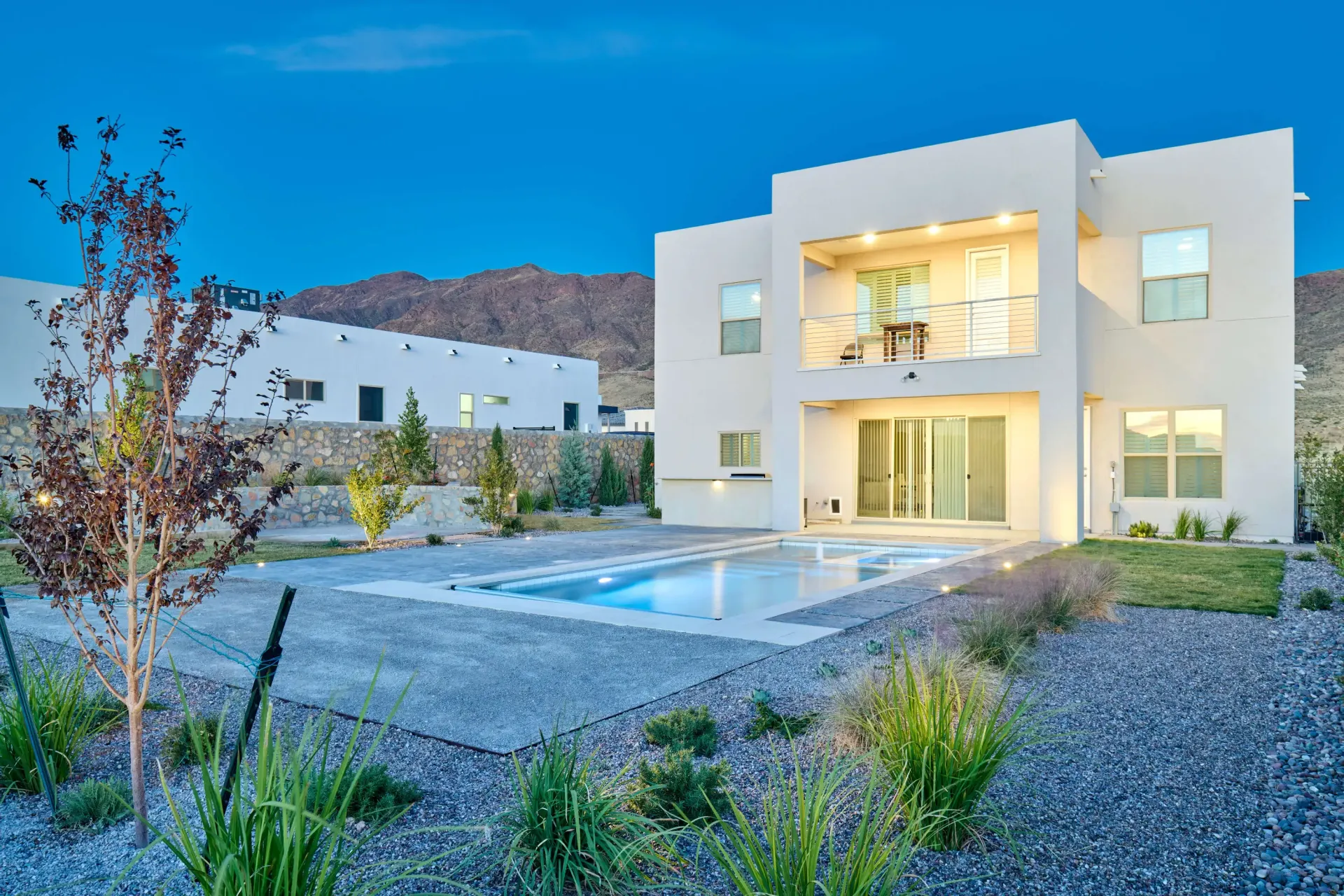 Modern two-story house with a pool in a desert landscape at dusk. Mountain in the background.