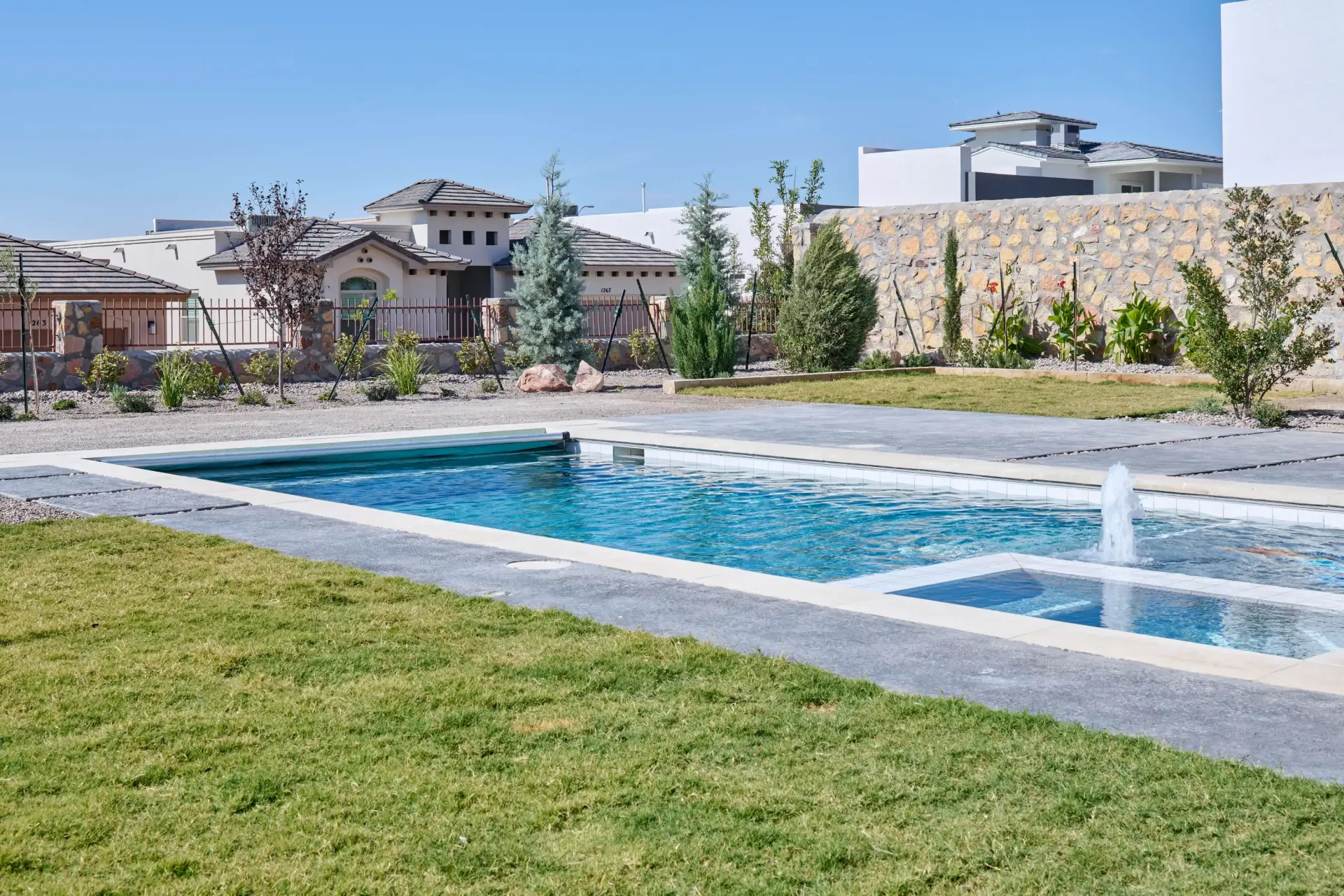Pool in a yard with a fountain, green grass, stone wall, and houses in the background on a sunny day.