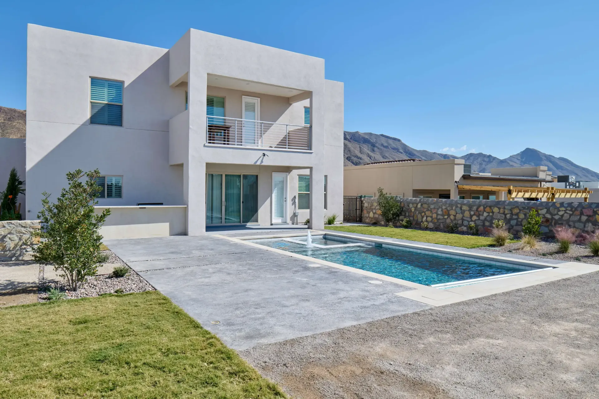 Modern two-story house with balcony, rectangular pool, and mountain backdrop under a clear blue sky.