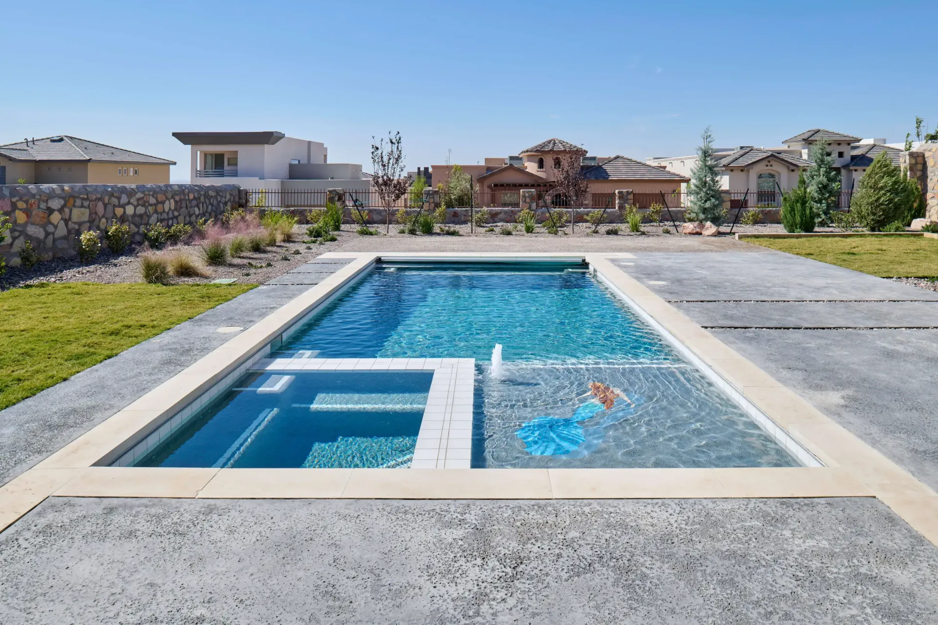 Rectangular swimming pool with attached spa, surrounded by concrete patio, in front of houses under a blue sky.