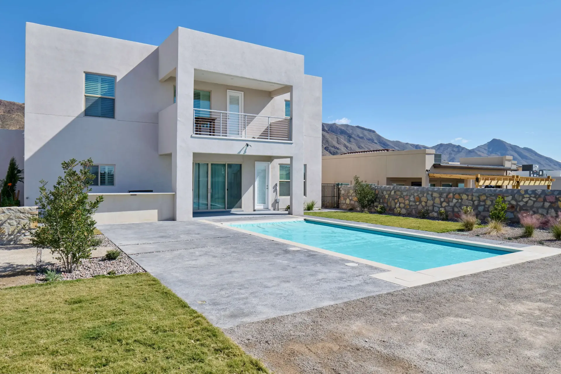 Modern two-story white house with a pool, patio, and balcony under a blue sky, mountains in the background.