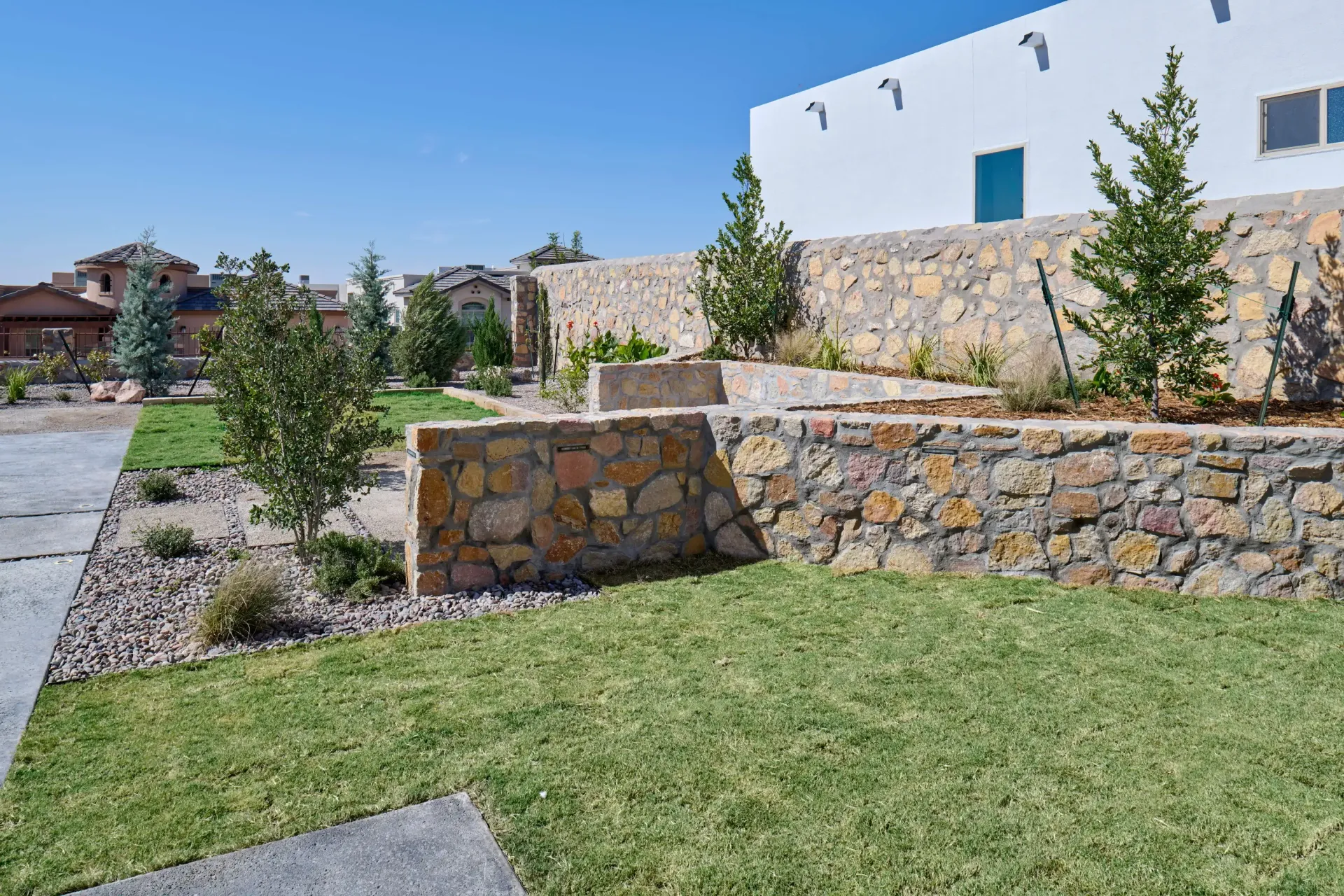 Landscaped yard with stone walls, grass, small trees, and a white building under a blue sky.