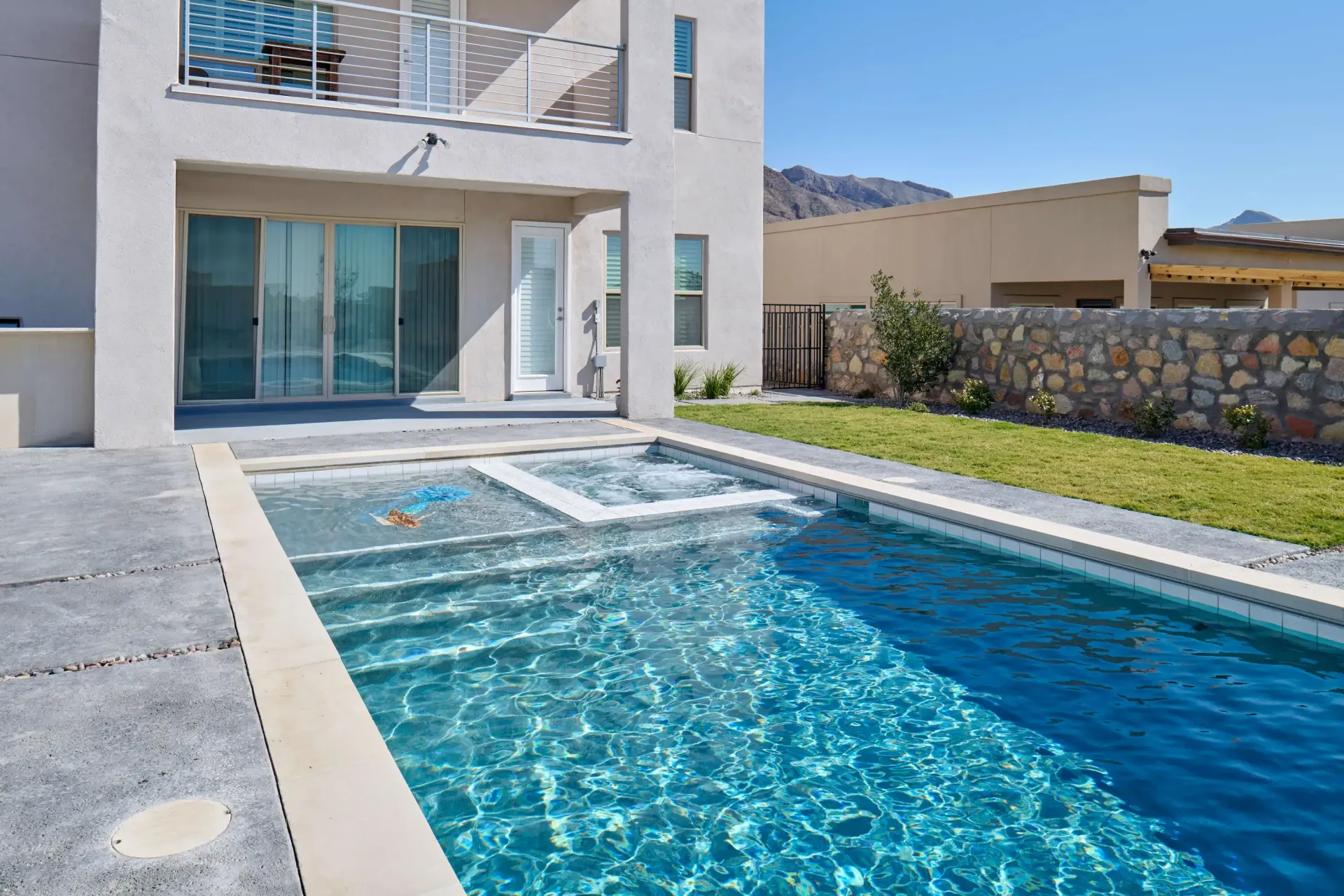 Exterior of a house with a pool and spa. Blue water, concrete patio, green lawn, and mountains in the distance.