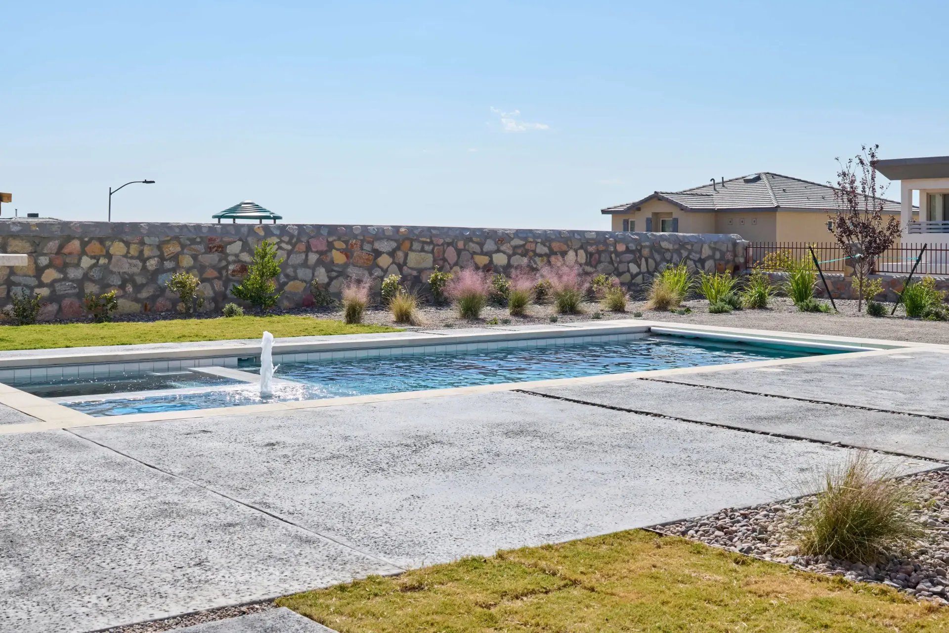 Rectangular pool with fountain, surrounded by concrete patio and stone wall, under a clear sky.