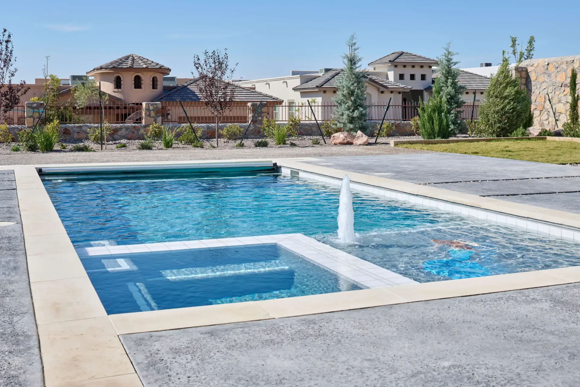 Swimming pool with fountain and spa, surrounded by concrete patio and landscaping, houses in the background.