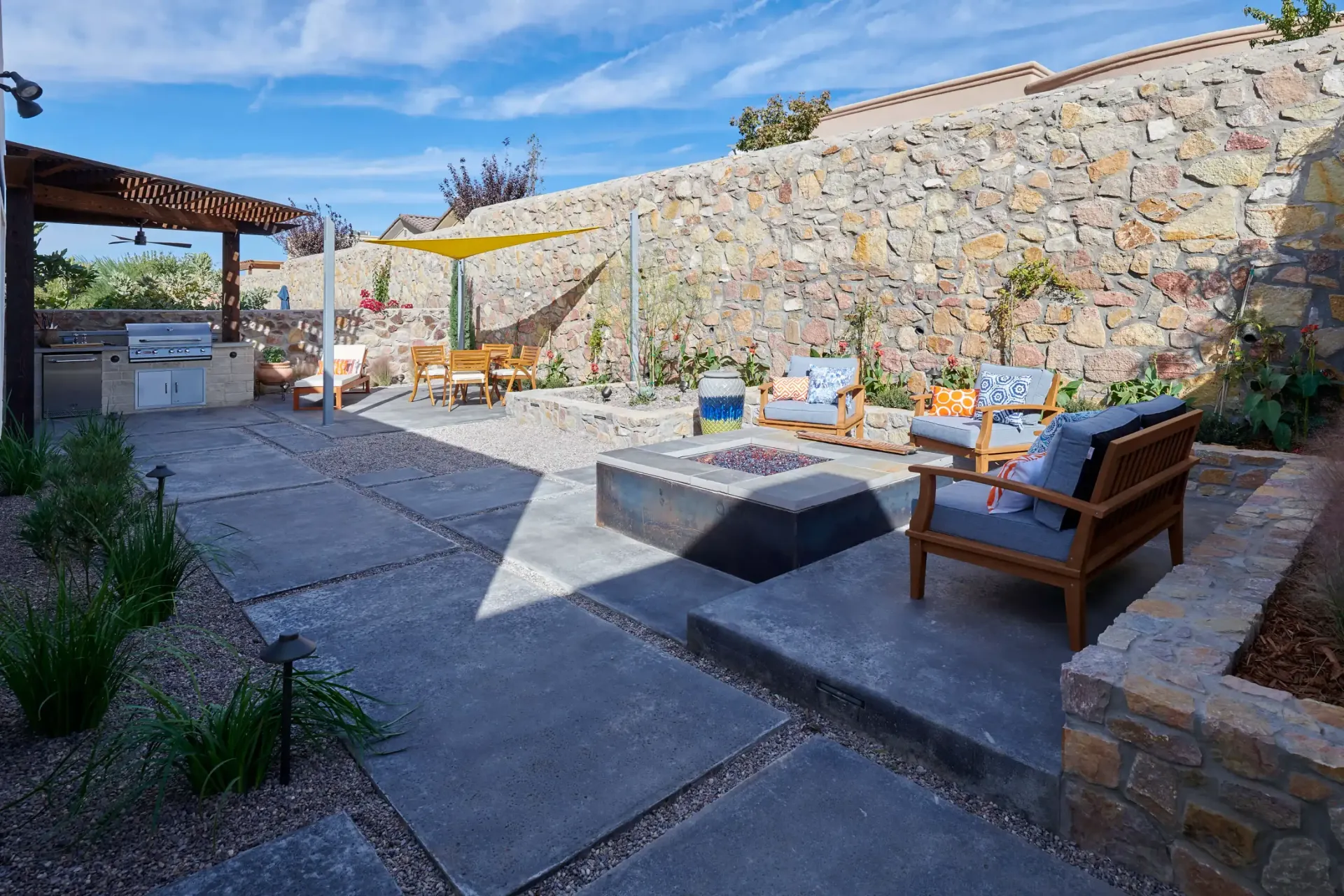 Outdoor patio with fireplace, seating, and built-in grill area, with a stone retaining wall and blue sky.