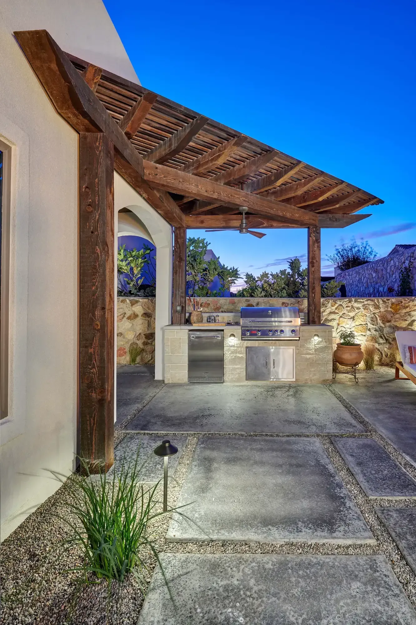 Outdoor kitchen with wooden pergola, grill, refrigerator, and stone patio.