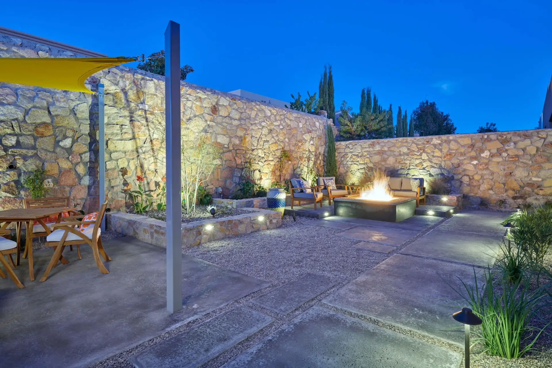 Stone-walled patio at dusk with fire pit, seating, and yellow shade. Uplighting illuminates the walls and plants.