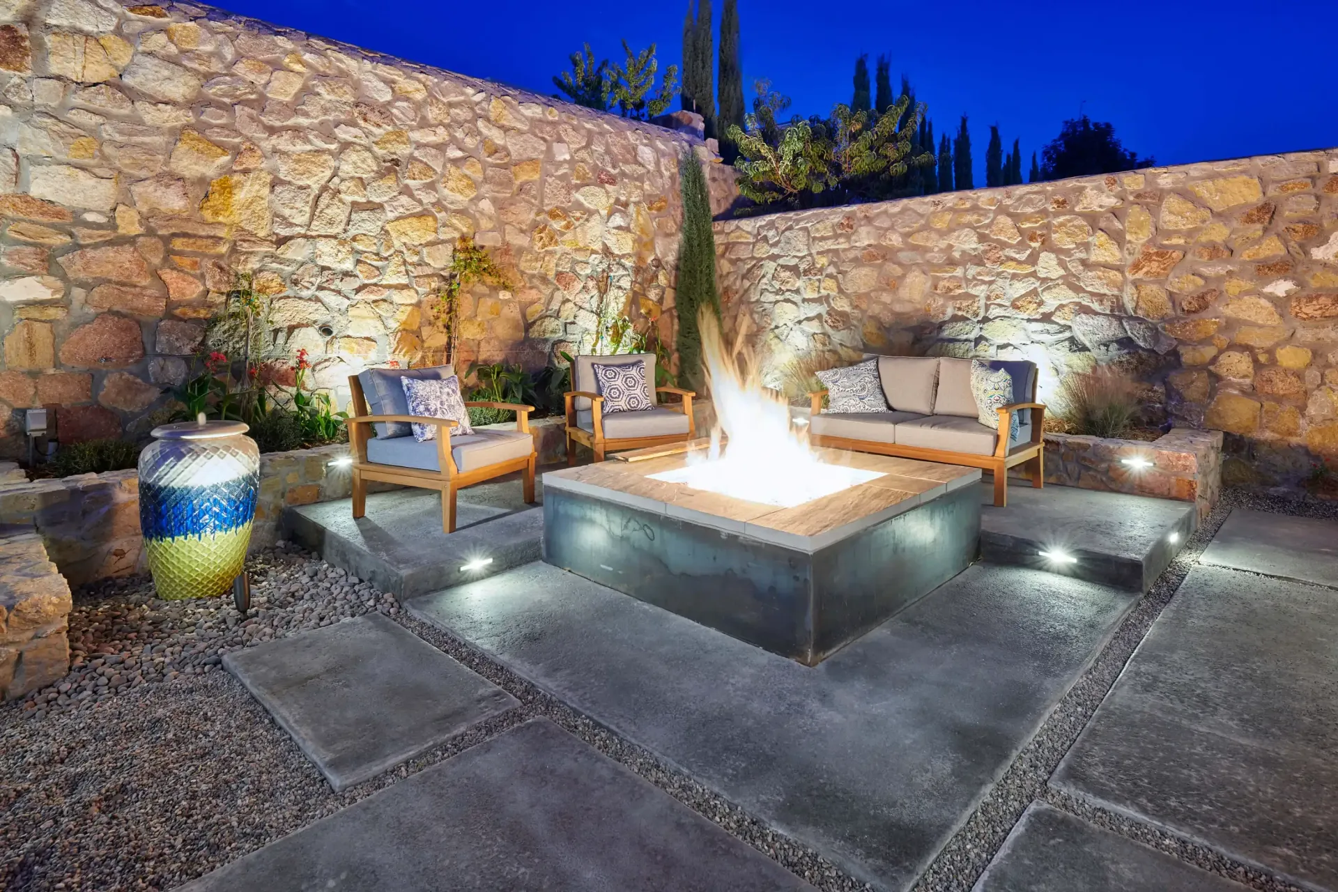 Outdoor seating area with lit fire pit and stone walls at dusk.