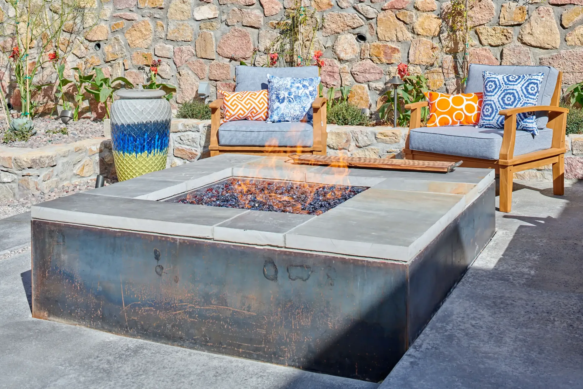 Outdoor fire pit with seating, stone wall background. Gray cushions, colorful pillows.