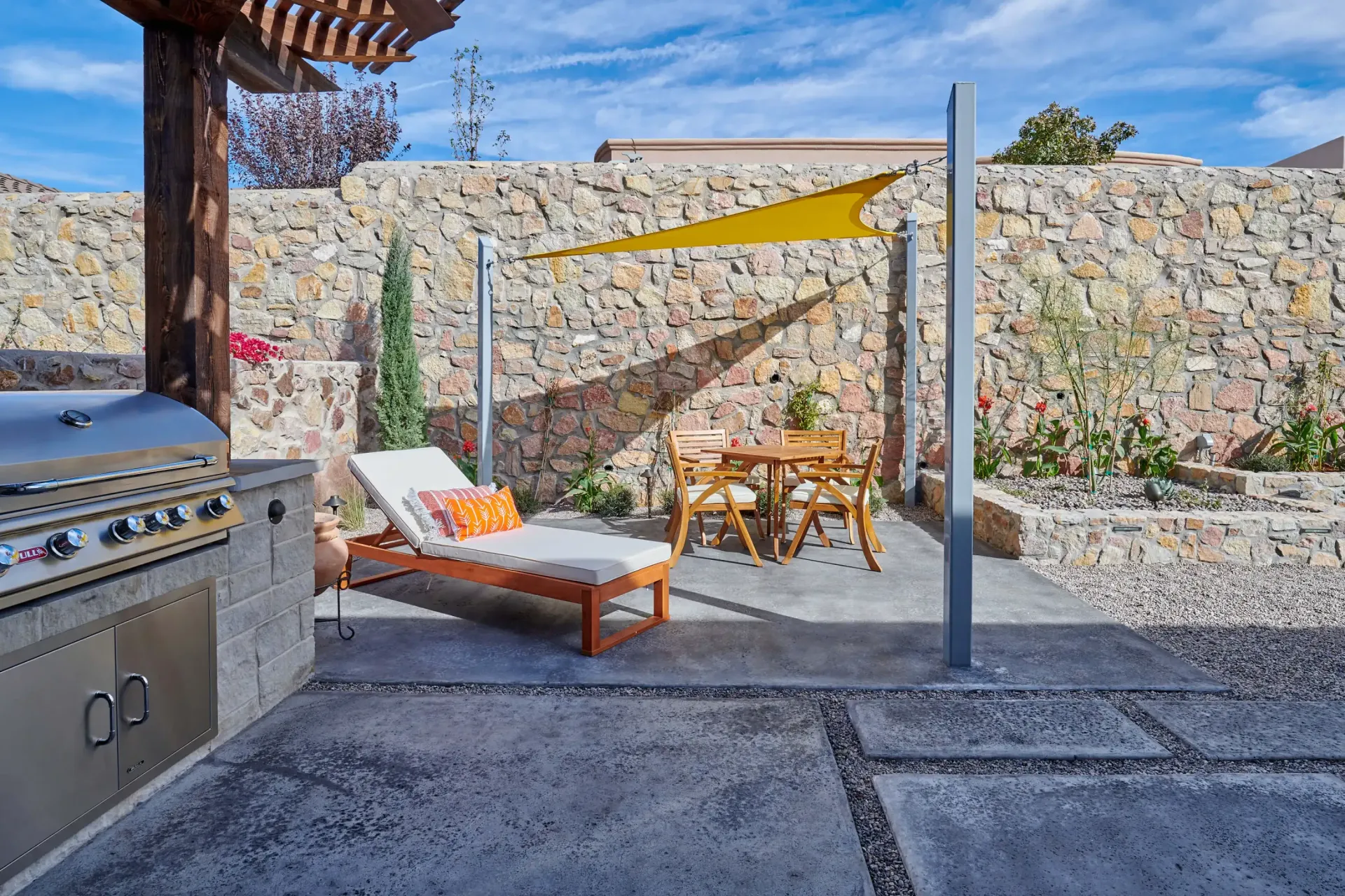 Outdoor patio with grill, lounge chair, table and shade sail against stone wall.