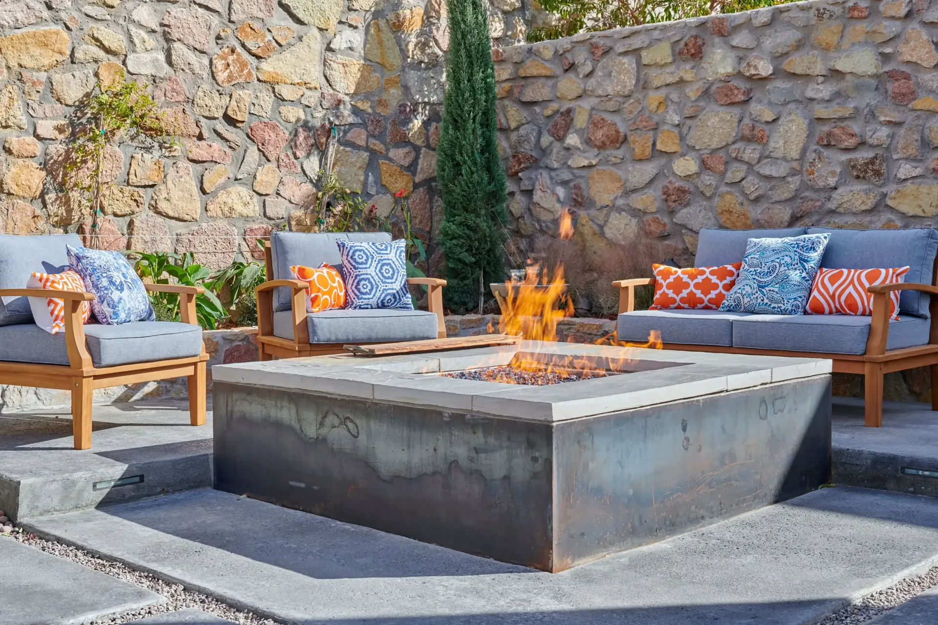 Outdoor seating area with lit fire pit and stone wall backdrop.