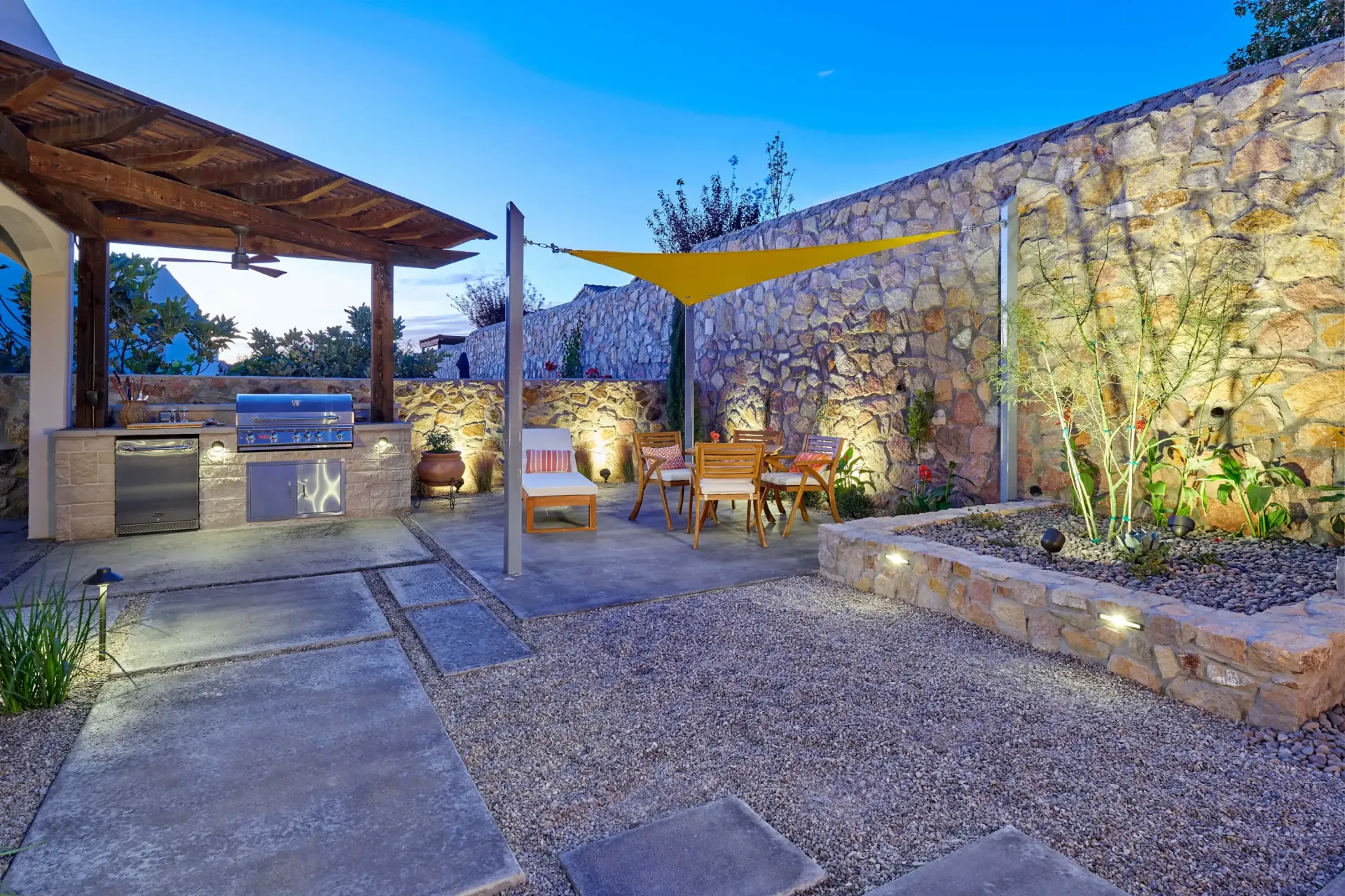 Outdoor patio with grill, seating, stone walls, and gravel ground. Yellow sun shade.