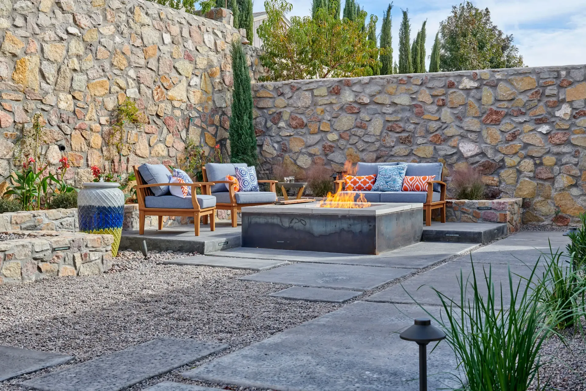 Outdoor patio with seating around a fire pit, bordered by stone walls and gravel pathways.