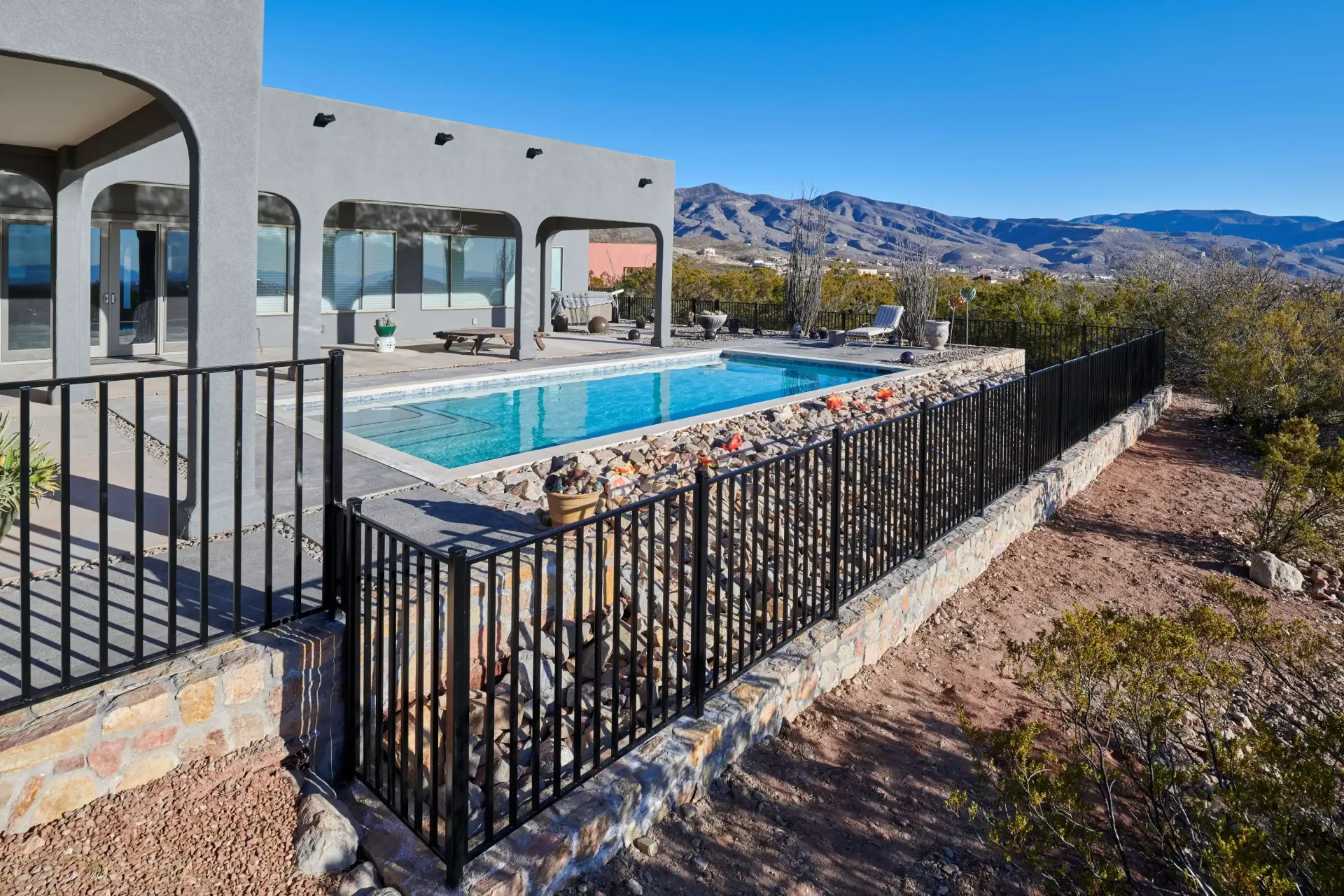Black fenced pool, stone wall, and desert landscape with house under clear blue sky.