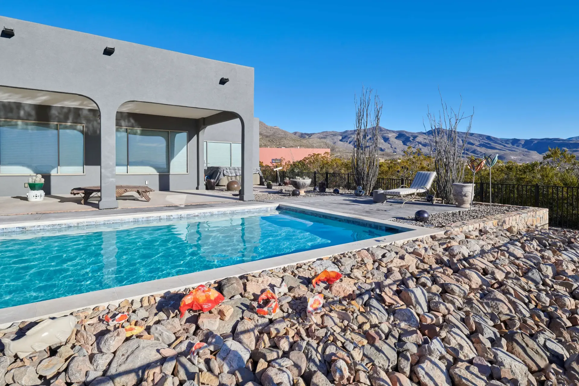 Poolside view: blue pool, stone retaining wall, grey stucco building, mountain backdrop.