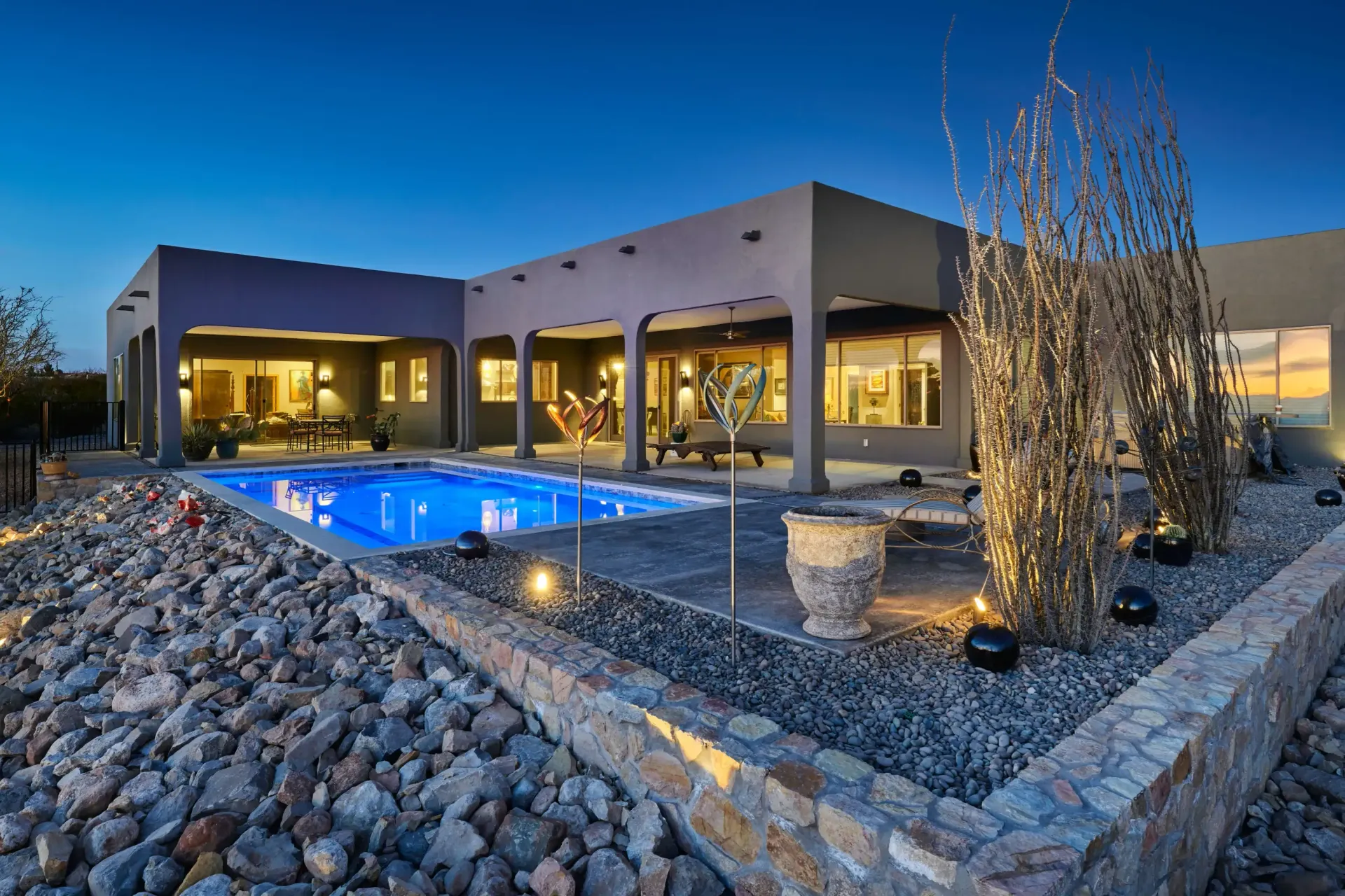 Modern home with a pool at dusk, lit patio, dark blue sky, gravel landscaping.