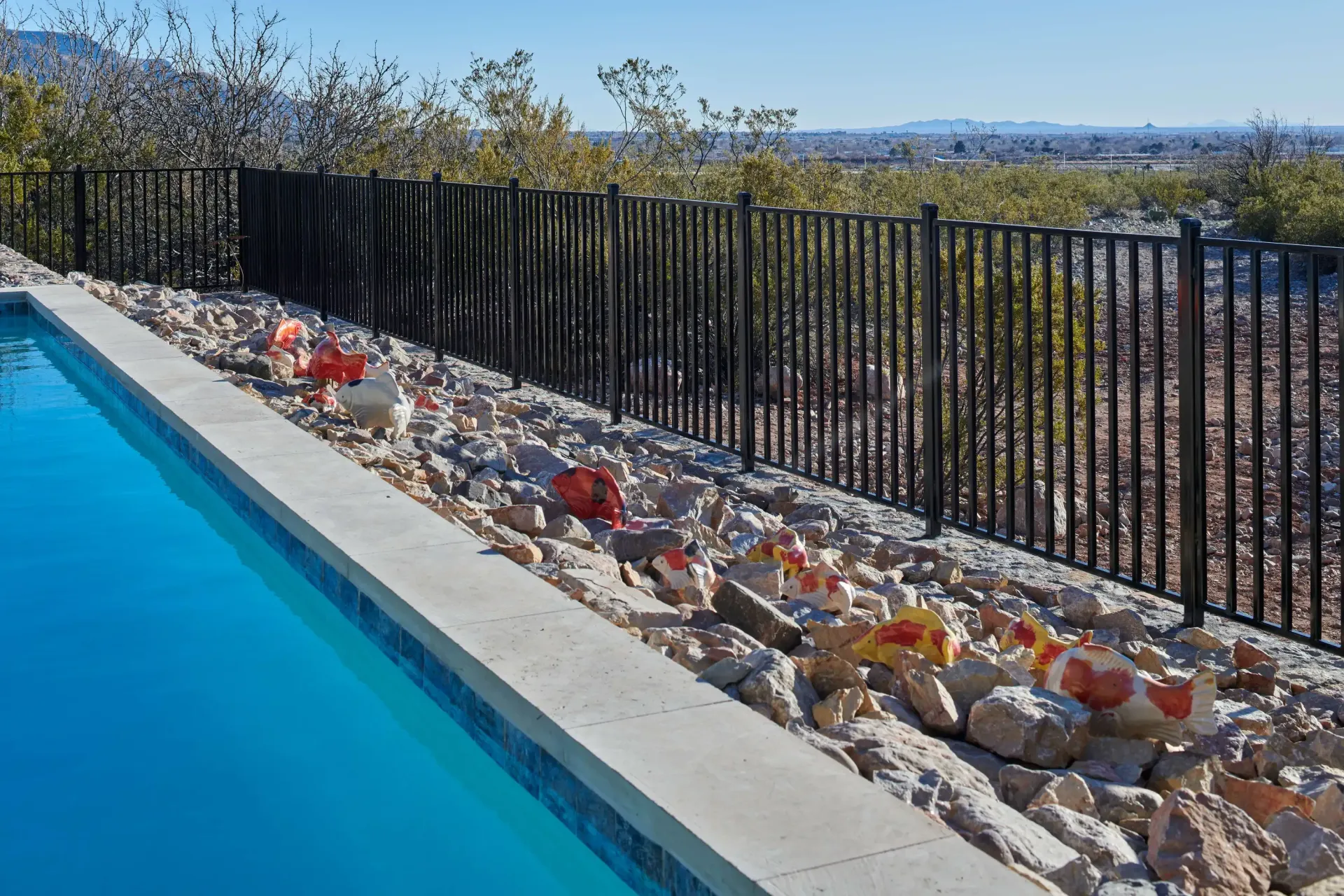 Poolside view: Black fence beside a pool with blue water, rocks, and desert vegetation under a blue sky.