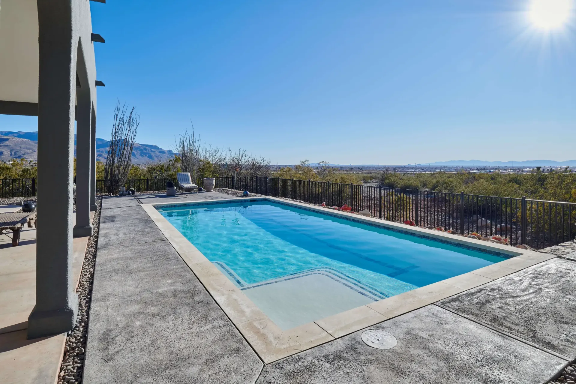 Rectangular pool on a concrete patio with a view of a town under a clear sky.