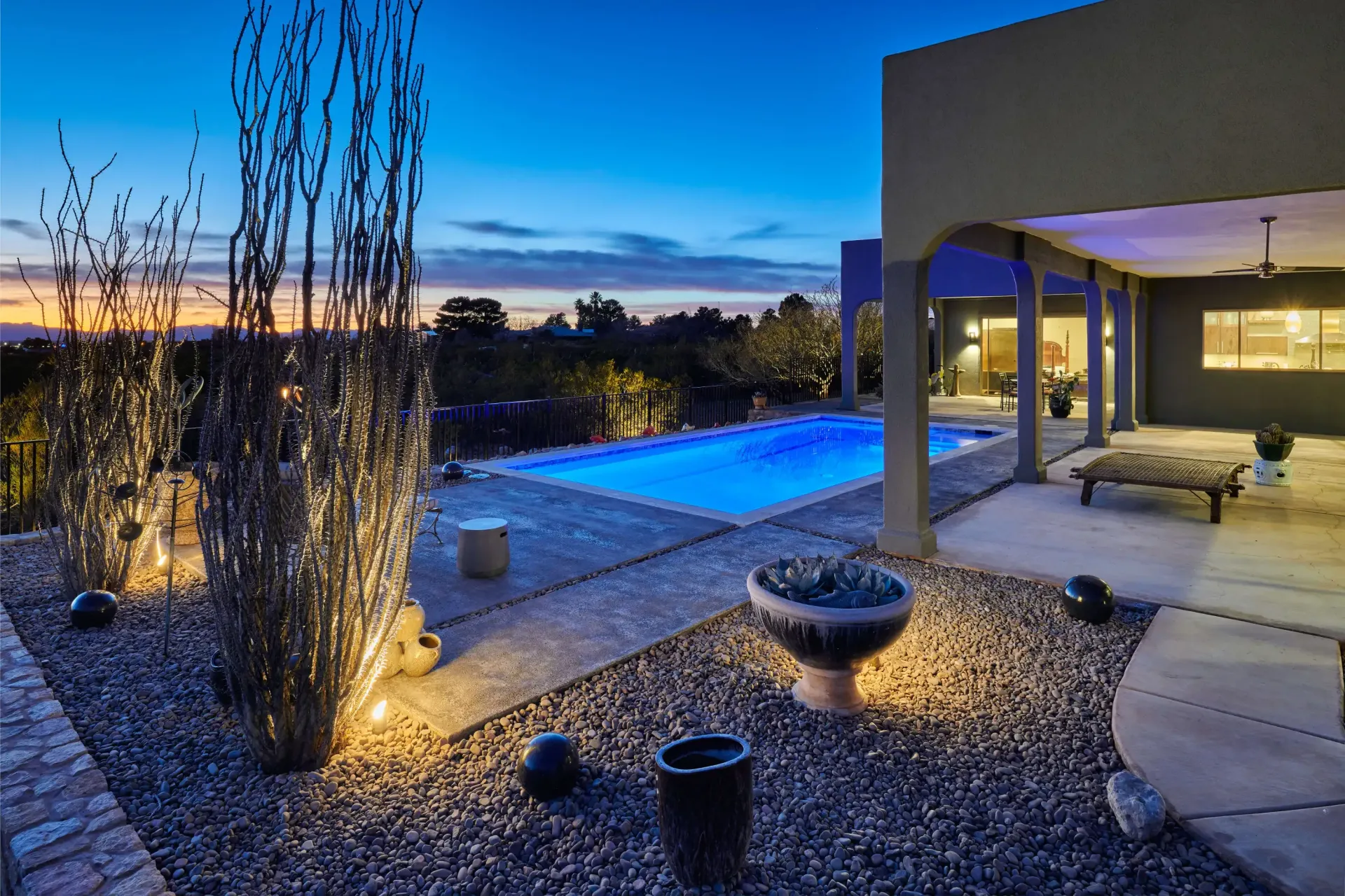 Night view of a backyard with a pool, patio, and illuminated landscaping against a twilight sky.