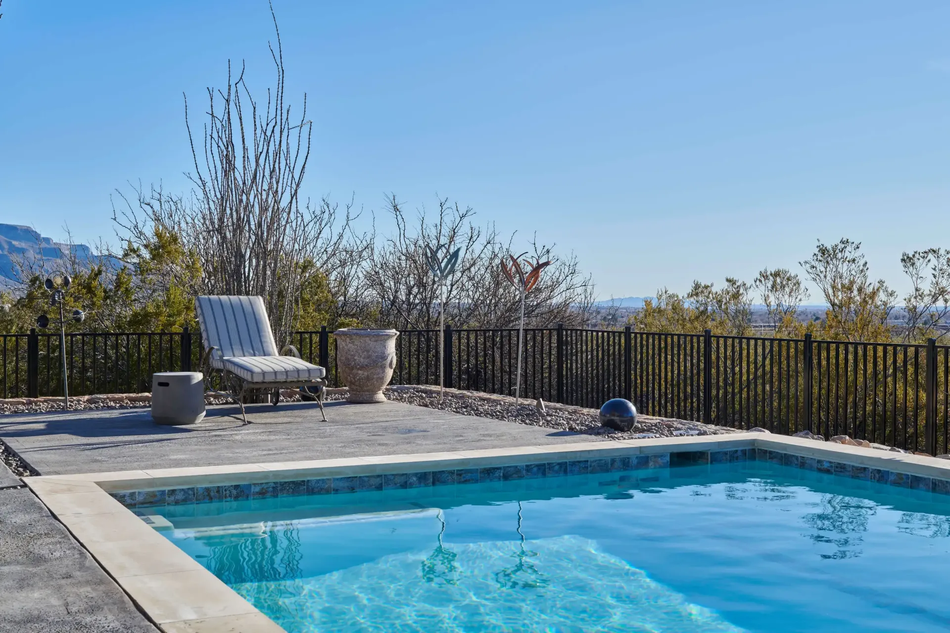 Poolside scene: blue pool, lounge chair, pot, black fence, and trees under a clear blue sky.
