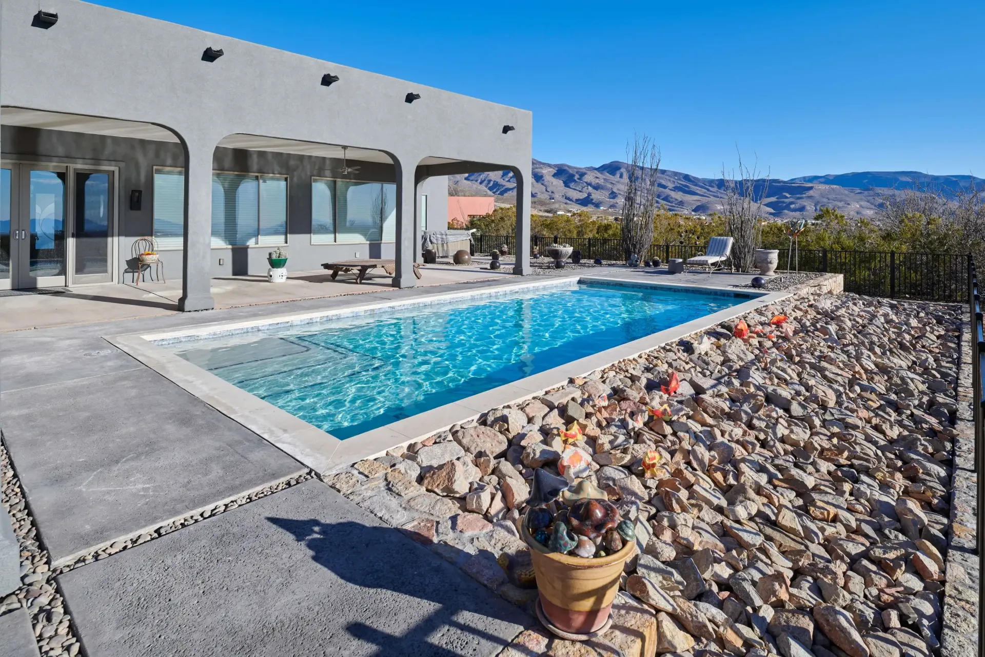 A rectangular pool next to a desert-landscaped yard, house with arches in the background, clear blue sky.