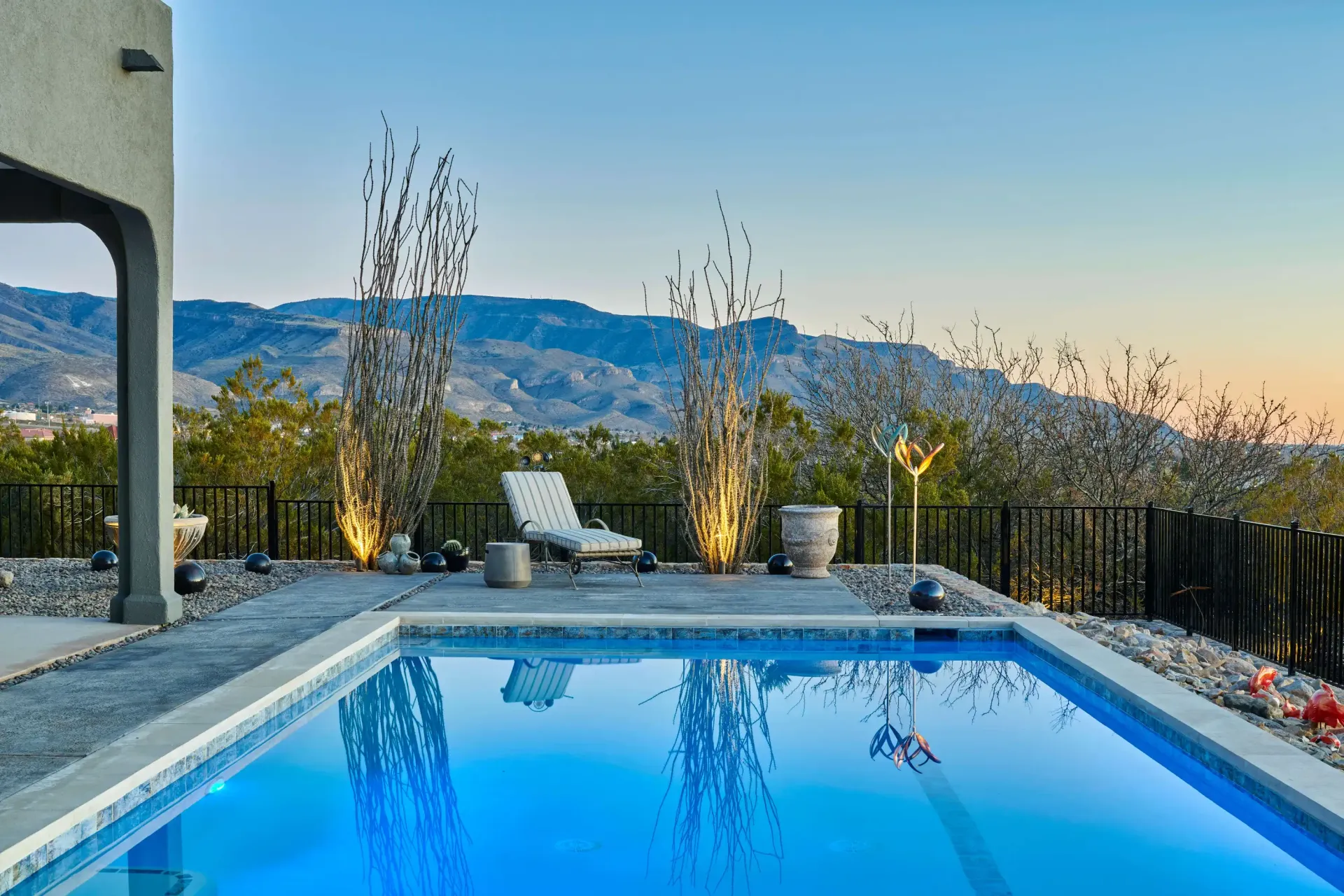 Rectangular pool with blue water overlooks mountains at dusk. Lounge chair and decorative trees on deck.