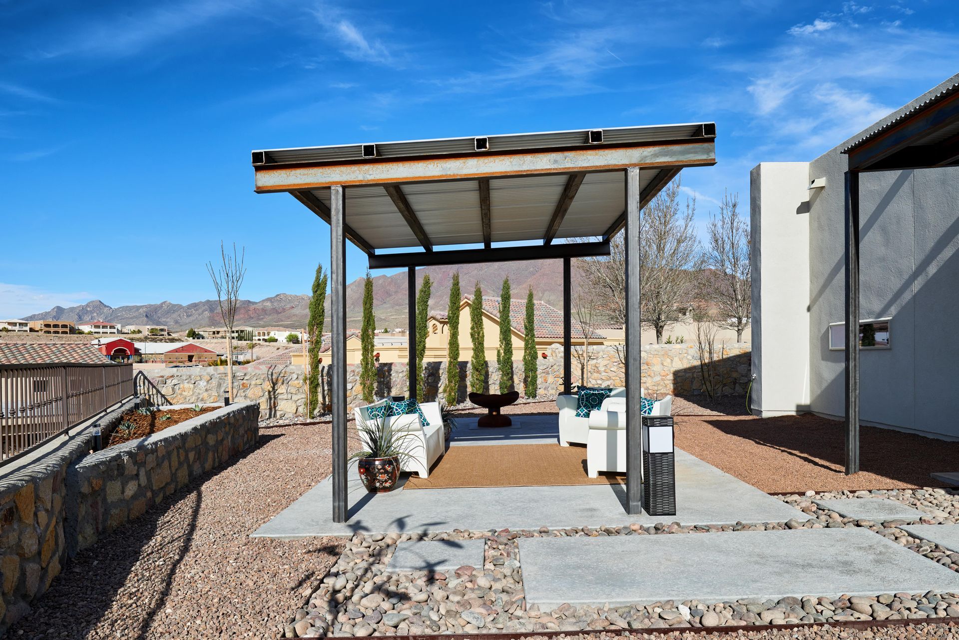 Outdoor seating area under a metal pergola; white furniture, blue cushions, green trees in background, sunny day.