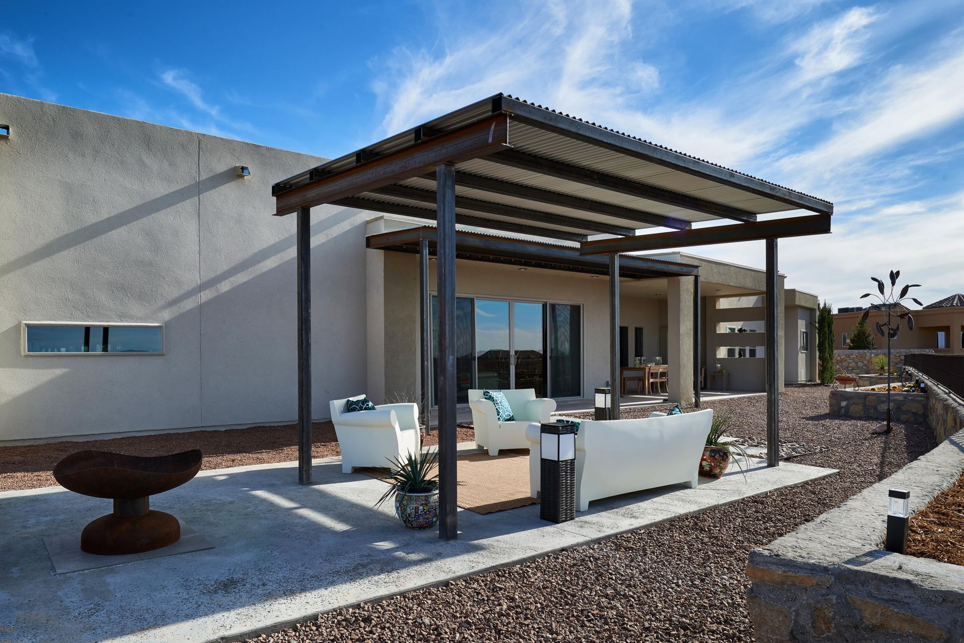 Patio with white furniture under a pergola, adjacent to a house with blue sky background.