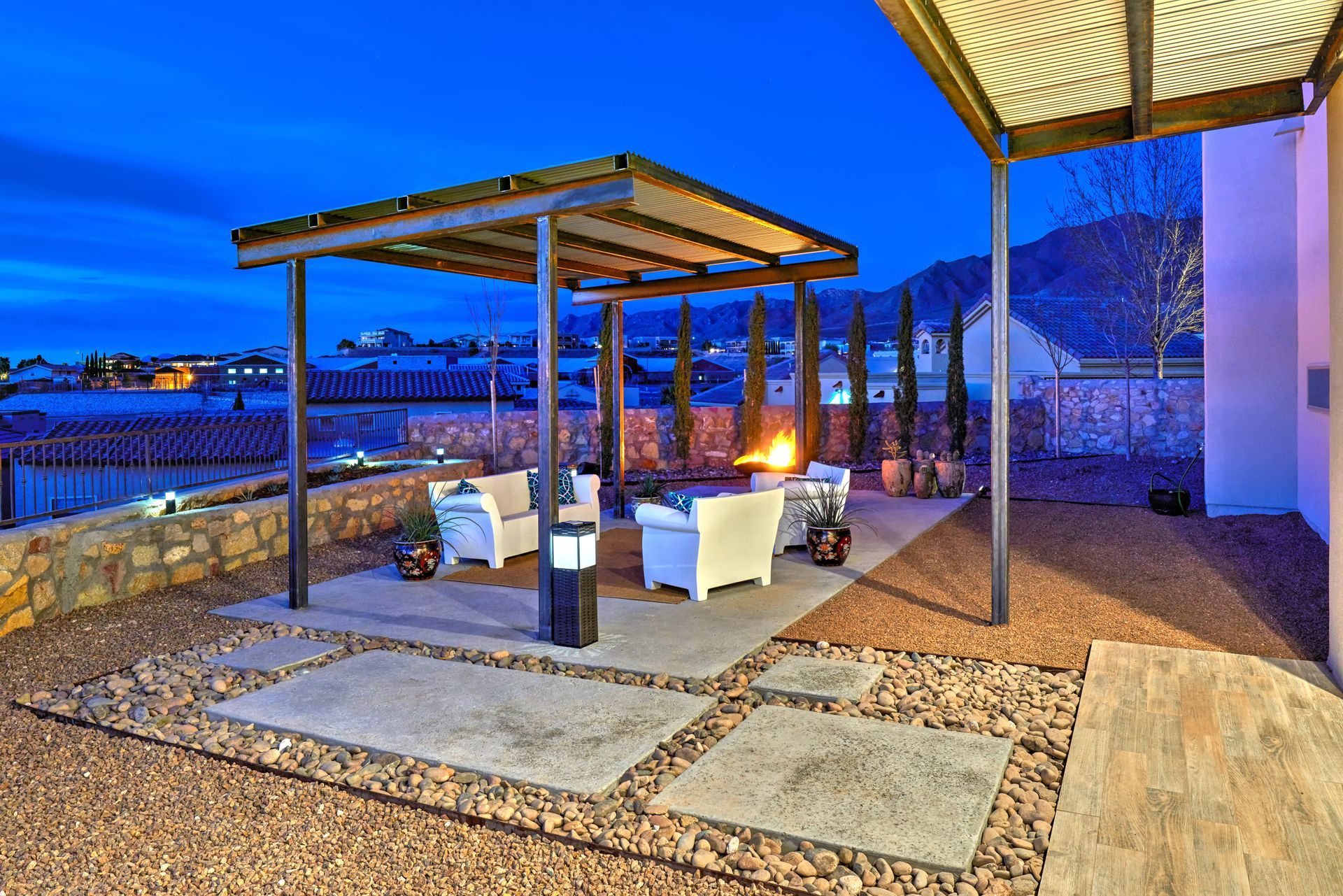 Outdoor patio with seating under a pergola, a fire pit, and gravel flooring at dusk.