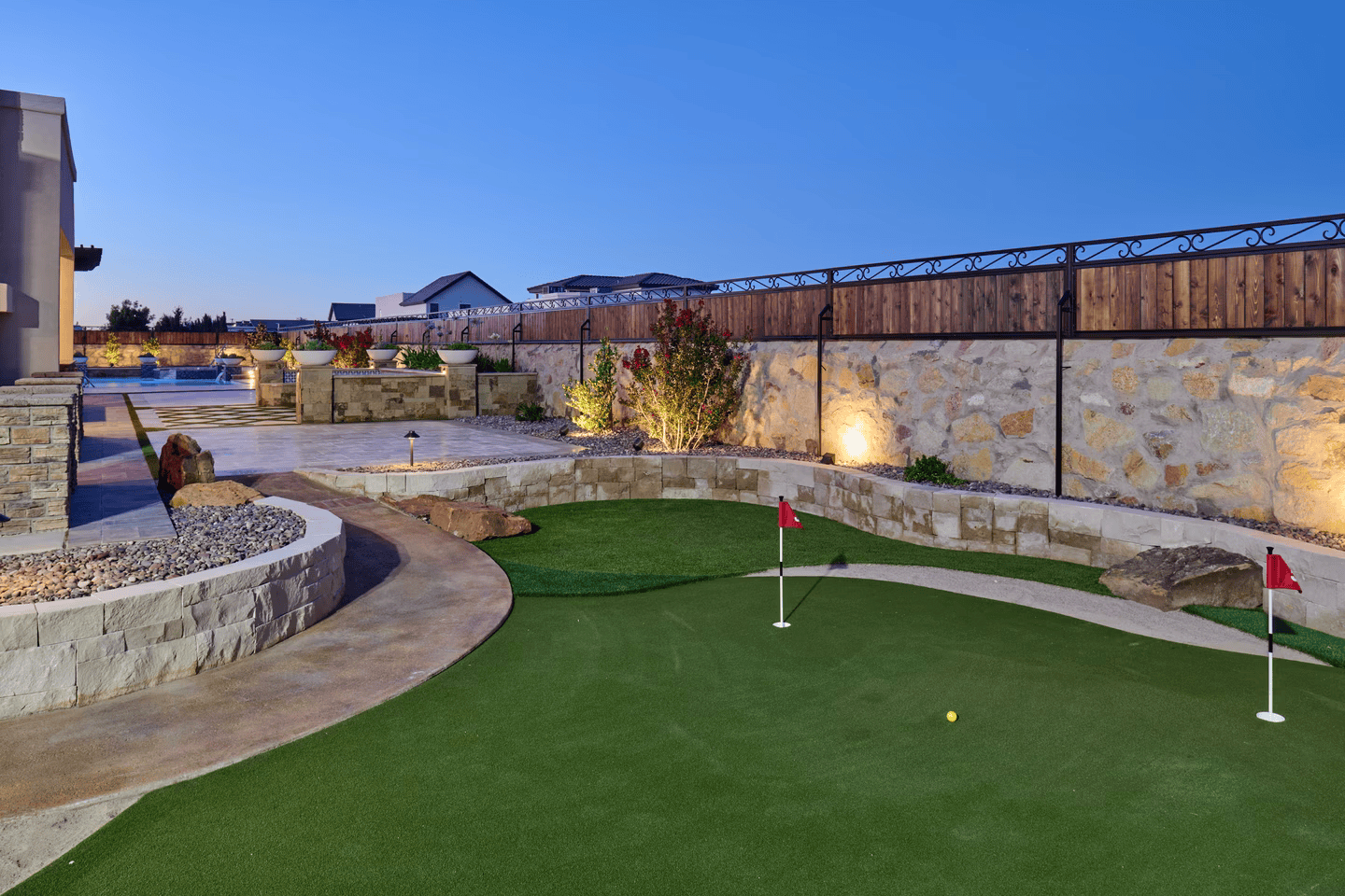 A backyard putting green with two holes, lit by spotlights, stone wall surrounds, and a wooden fence.
