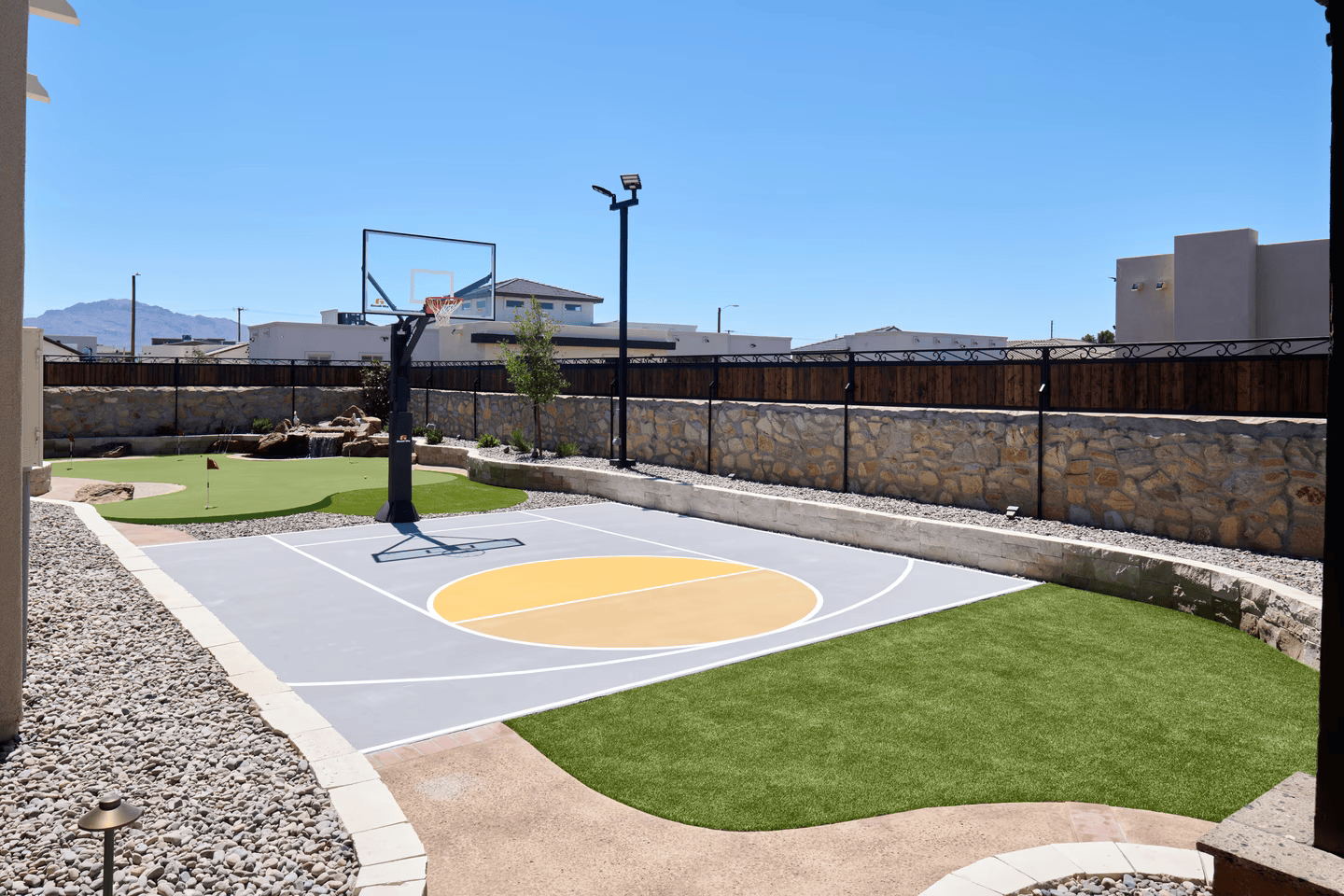 Backyard basketball court with a yellow center, green turf, and rock walls under a blue sky.